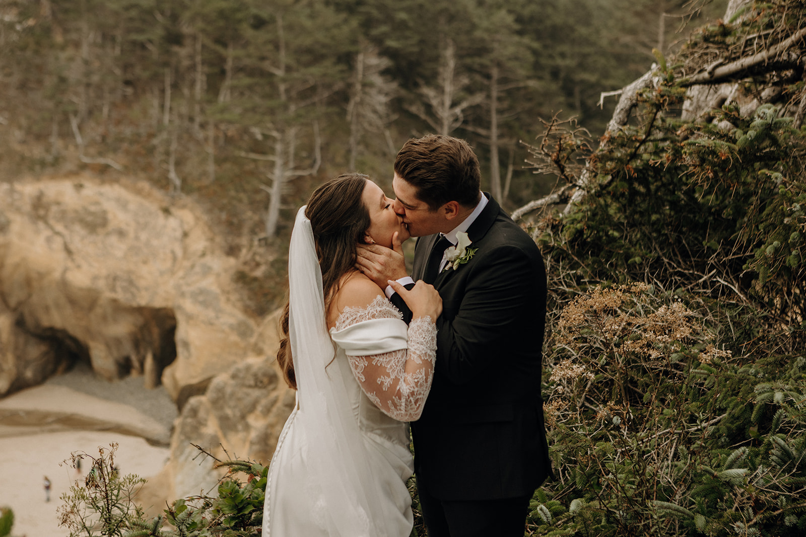 A couple in wedding attire shares a kiss on a cliffside, surrounded by lush greenery and overlooking a rocky beach, conveying romance and joy.