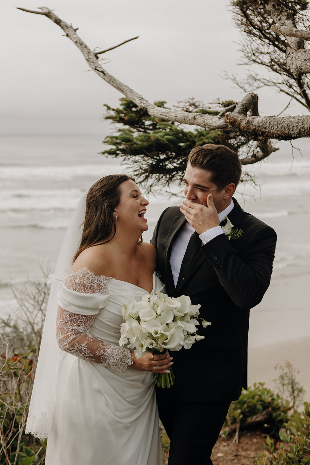 Bride and groom laughing joyfully by the beach, the bride holds white flowers, the groom covers his mouth. Windswept tree in the background.