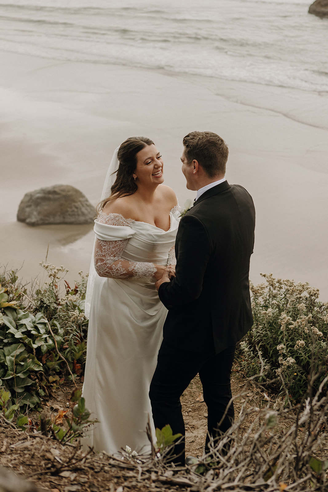 A bride in a white dress and veil and groom in a black suit stand smiling and holding hands on a sandy beach, with ocean waves in the background.