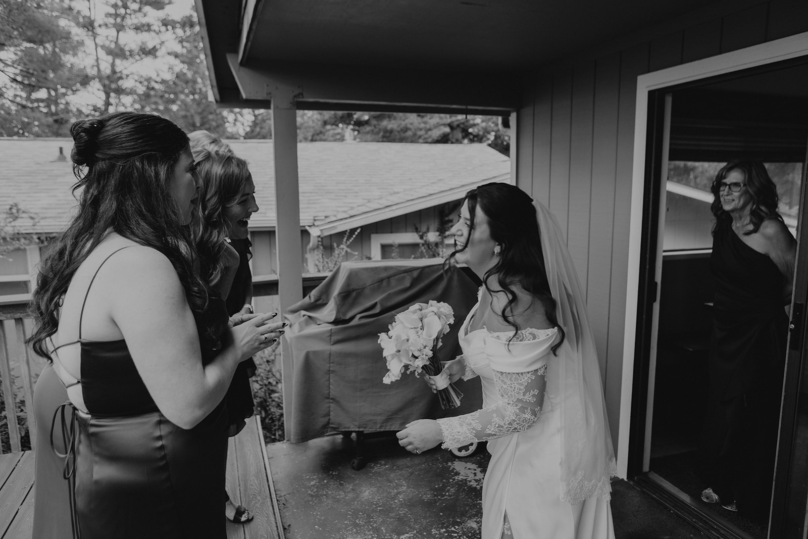 A bride in an elegant gown and veil, holding a bouquet, laughs with two women on a porch. Another woman watches from a doorway, smiling warmly.