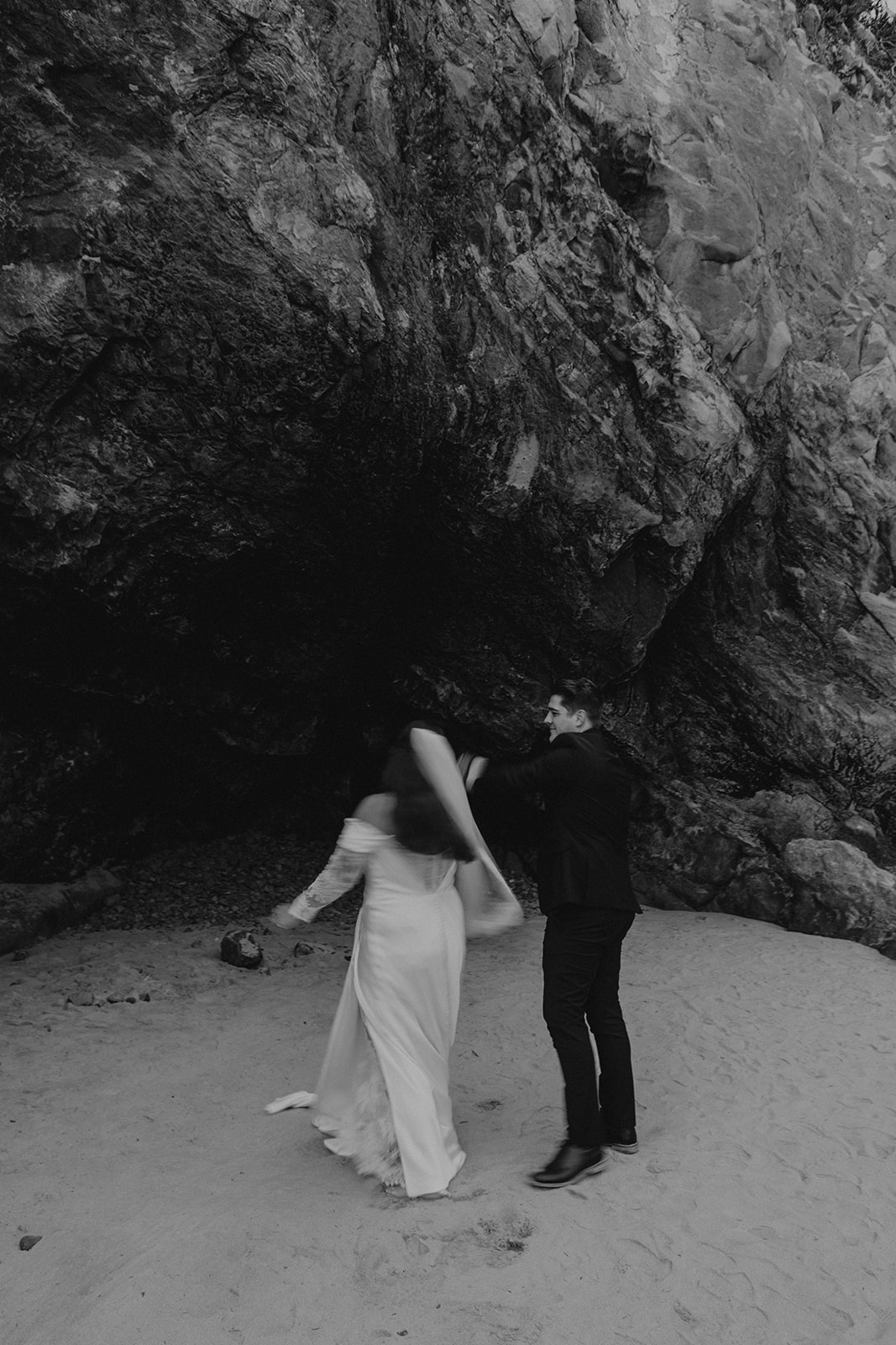 A black-and-white photo of a couple dancing joyfully on a sandy beach near a rocky cliff. The woman’s long dress sways elegantly, creating a lively scene.