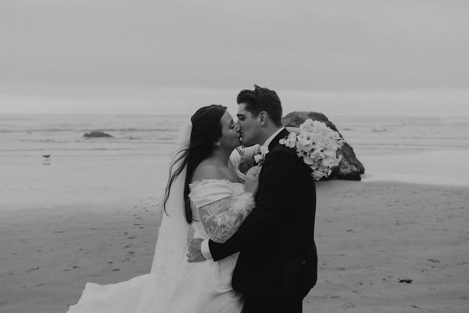 A couple in wedding attire shares a kiss on a serene beach. The bride holds a bouquet, and the sea creates a tranquil backdrop. Romantic and intimate.