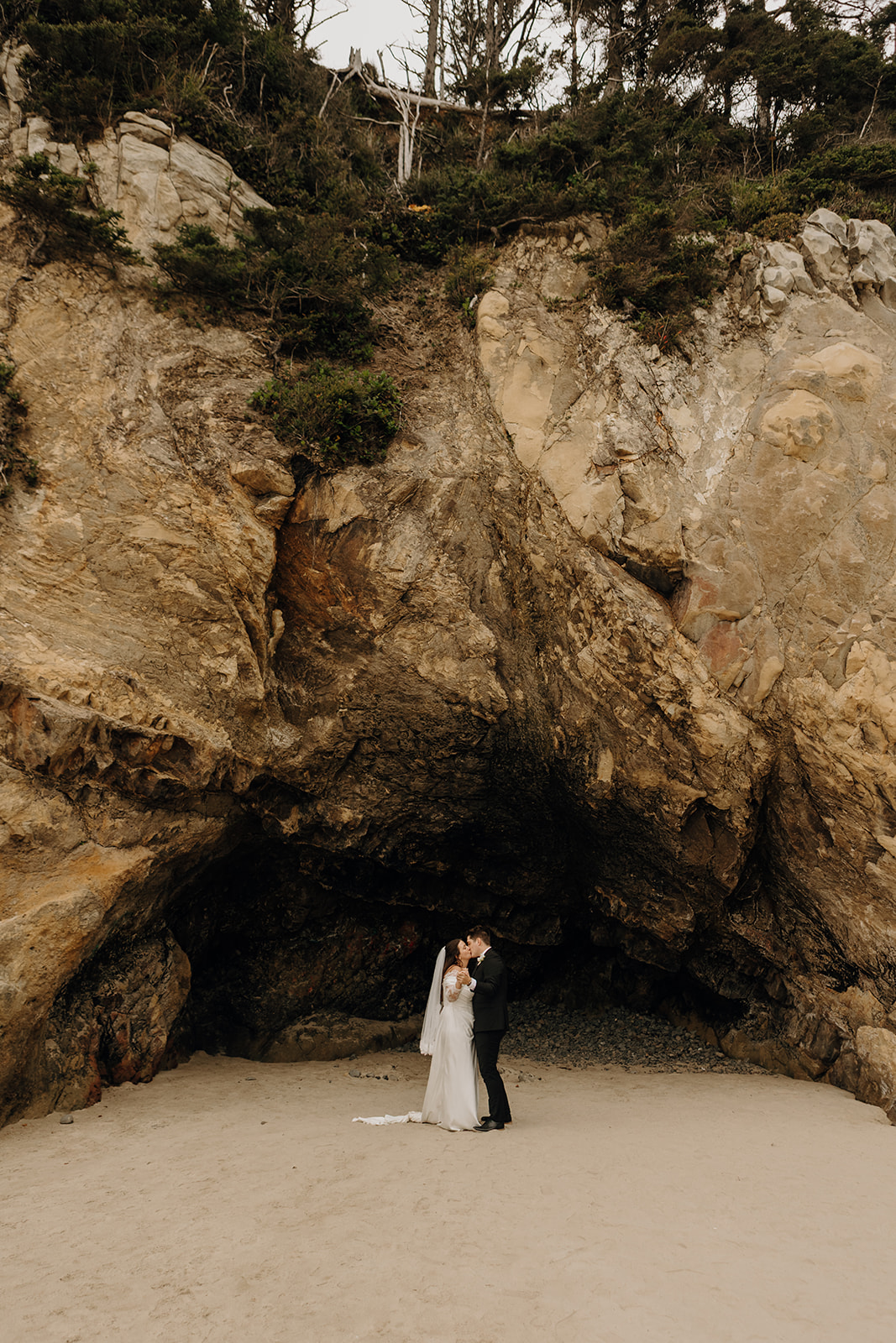 A couple embraces in a tender moment on a sandy beach, with a majestic rocky cave backdrop. The atmosphere is serene and romantic.