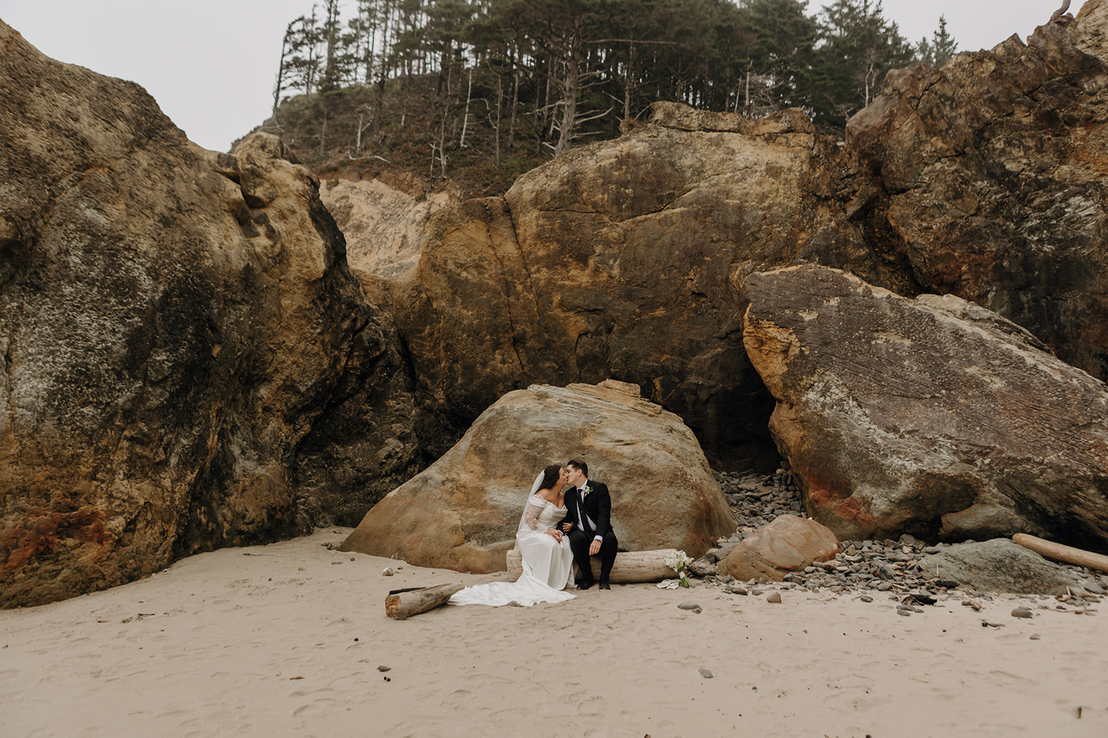 A bride in a white gown and groom in a black suit sit on a log, kissing in front of large, rugged boulders on a sandy beach near Cannon Beach, Oregon.