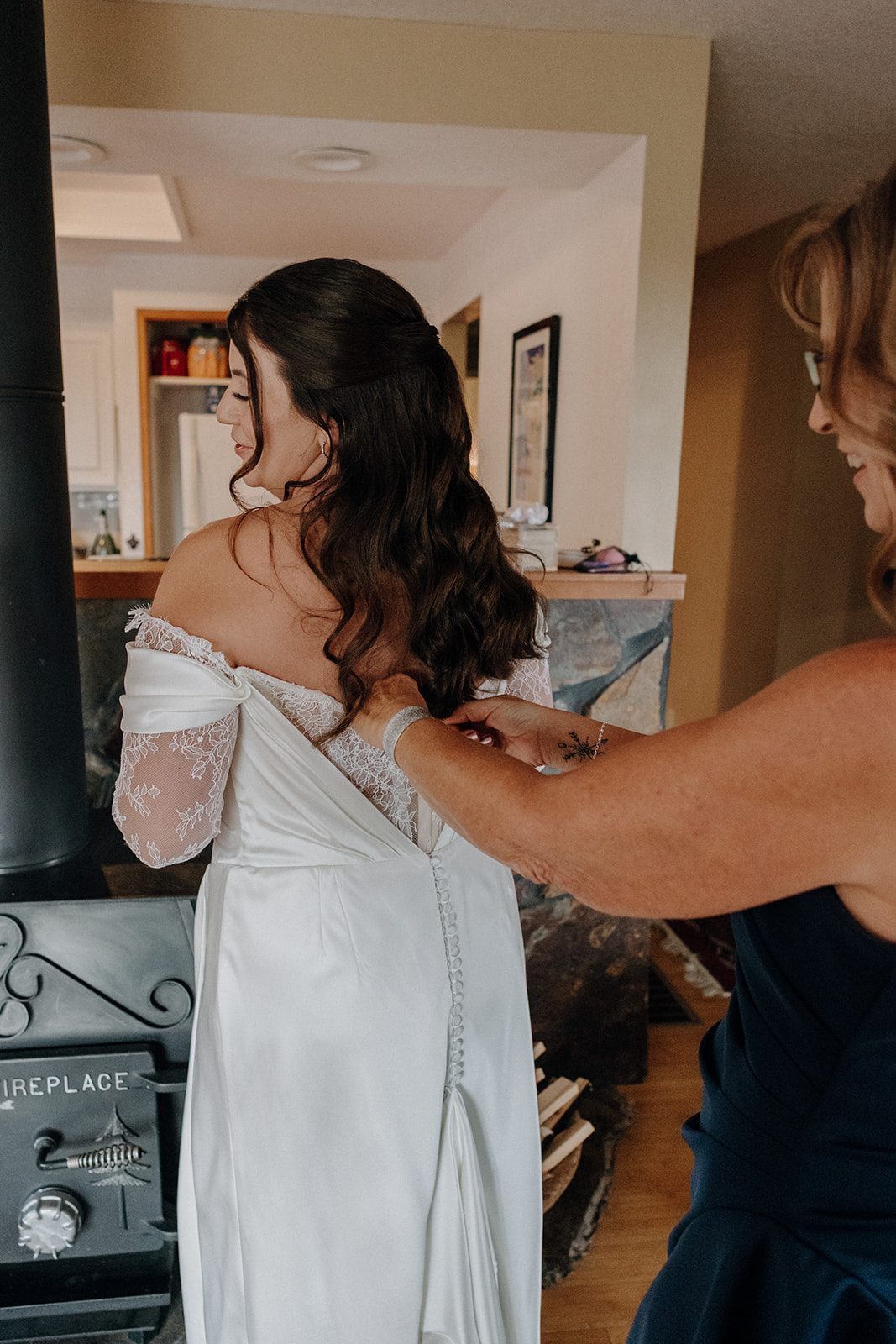 Bride in a white gown is having her dress zipped by an older woman. The room has a warm, cozy feel with visible kitchen elements and a fireplace.