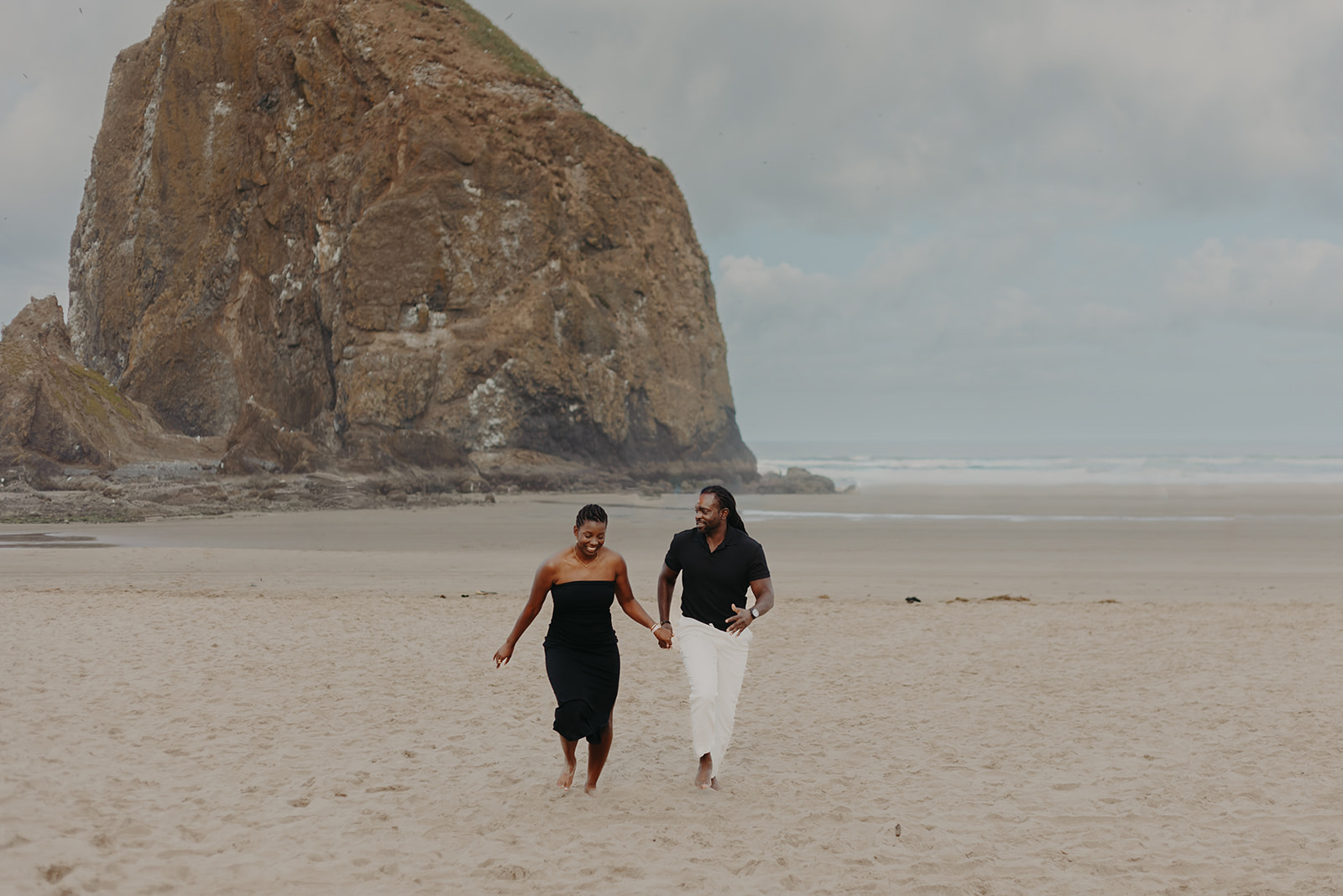 A couple walks hand in hand on a sandy beach, smiling. Haystack Rock rises in the background under a cloudy sky, conveying a relaxed and joyful mood.