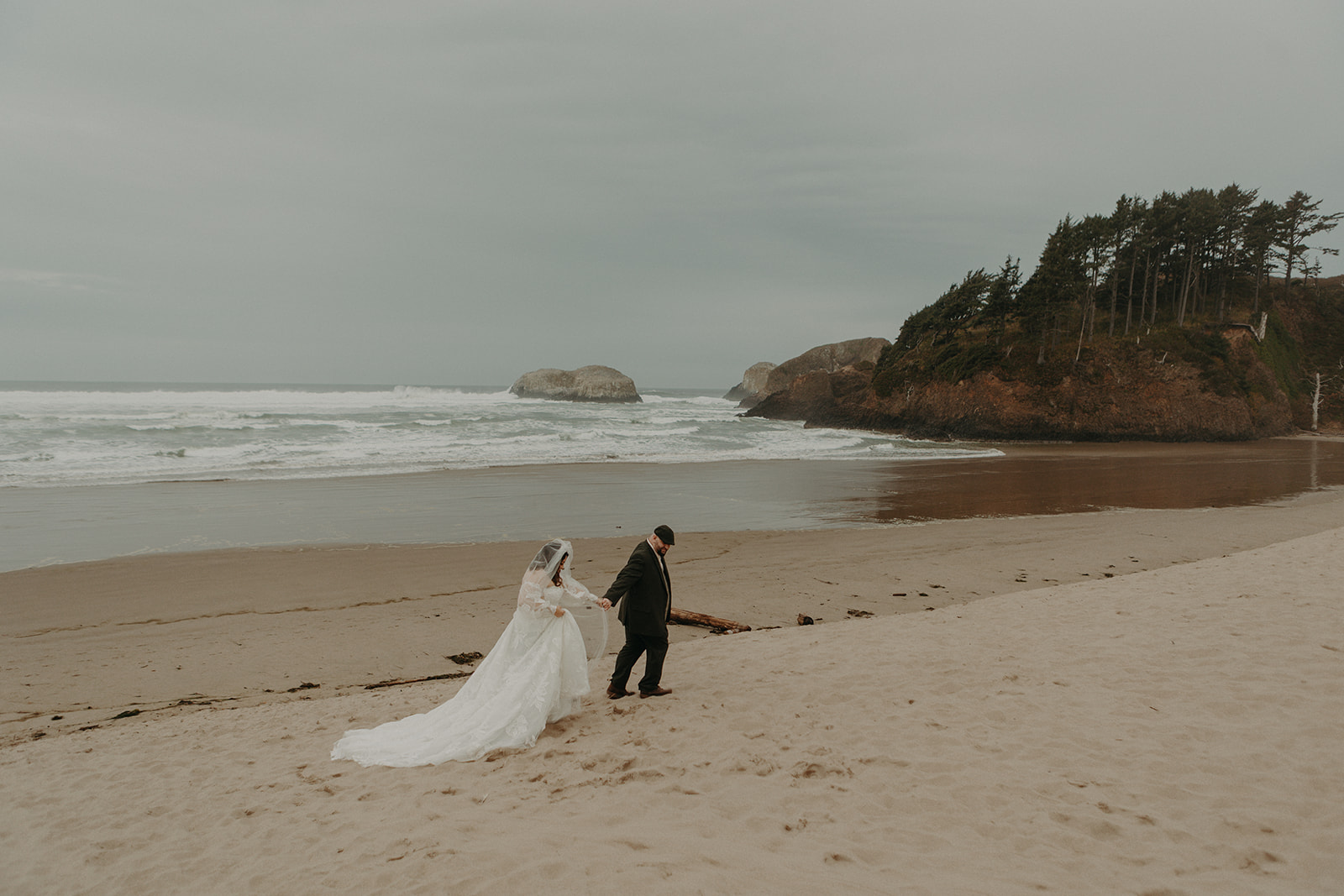A bride in a white dress and a groom in a suit walk hand-in-hand on Chapman beach at the Oregon Coast. Waves crash in the background under a cloudy sky, creating a romantic, tranquil scene.