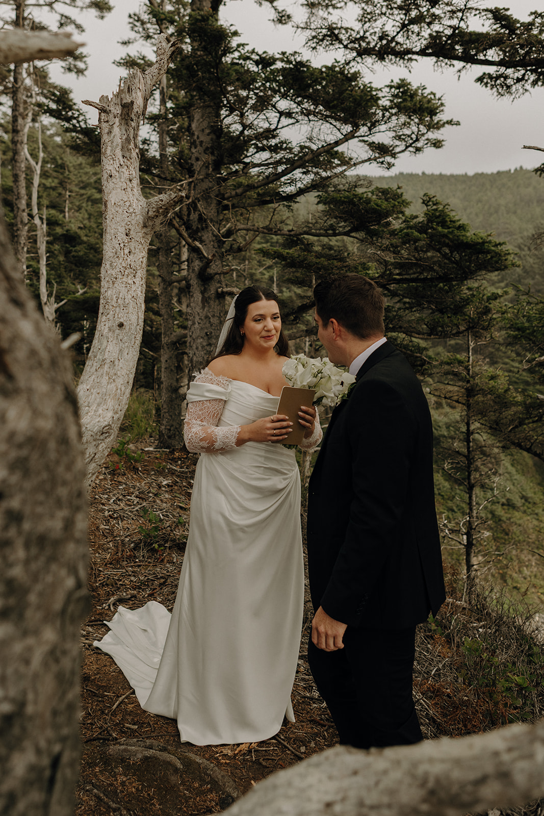 Bride in an elegant white gown reads vows to groom in a suit, standing in a serene forest setting. Emotionally intimate and romantic mood.