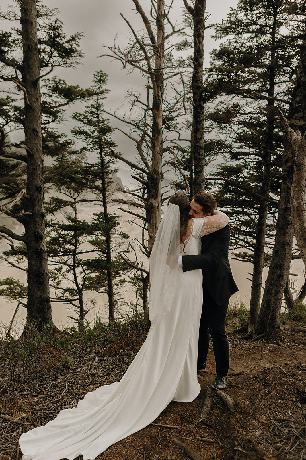 A couple embraces on a forested cliffside, the bride in a flowing white dress and veil. Tall trees and a misty ocean view create a serene, romantic atmosphere.
