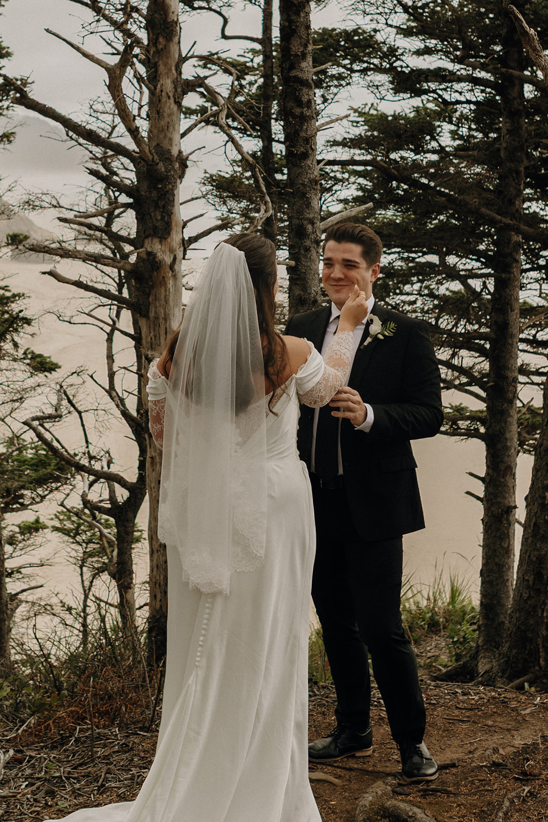 A bride in a white dress and veil gently touches the smiling groom's face in a forest setting. The couple stands among tall trees near the beach, conveying romance and serenity.
