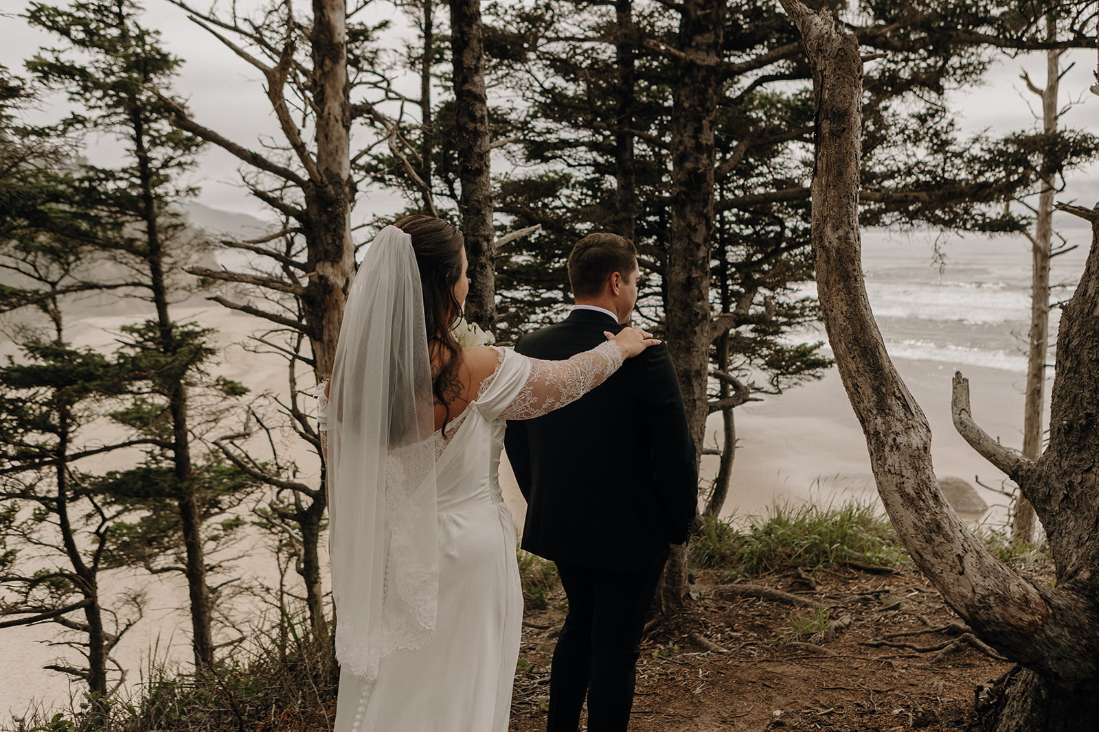 Bride in white gown and veil gently touches groom's shoulder from behind. They stand amidst trees overlooking a serene, cloudy beach.