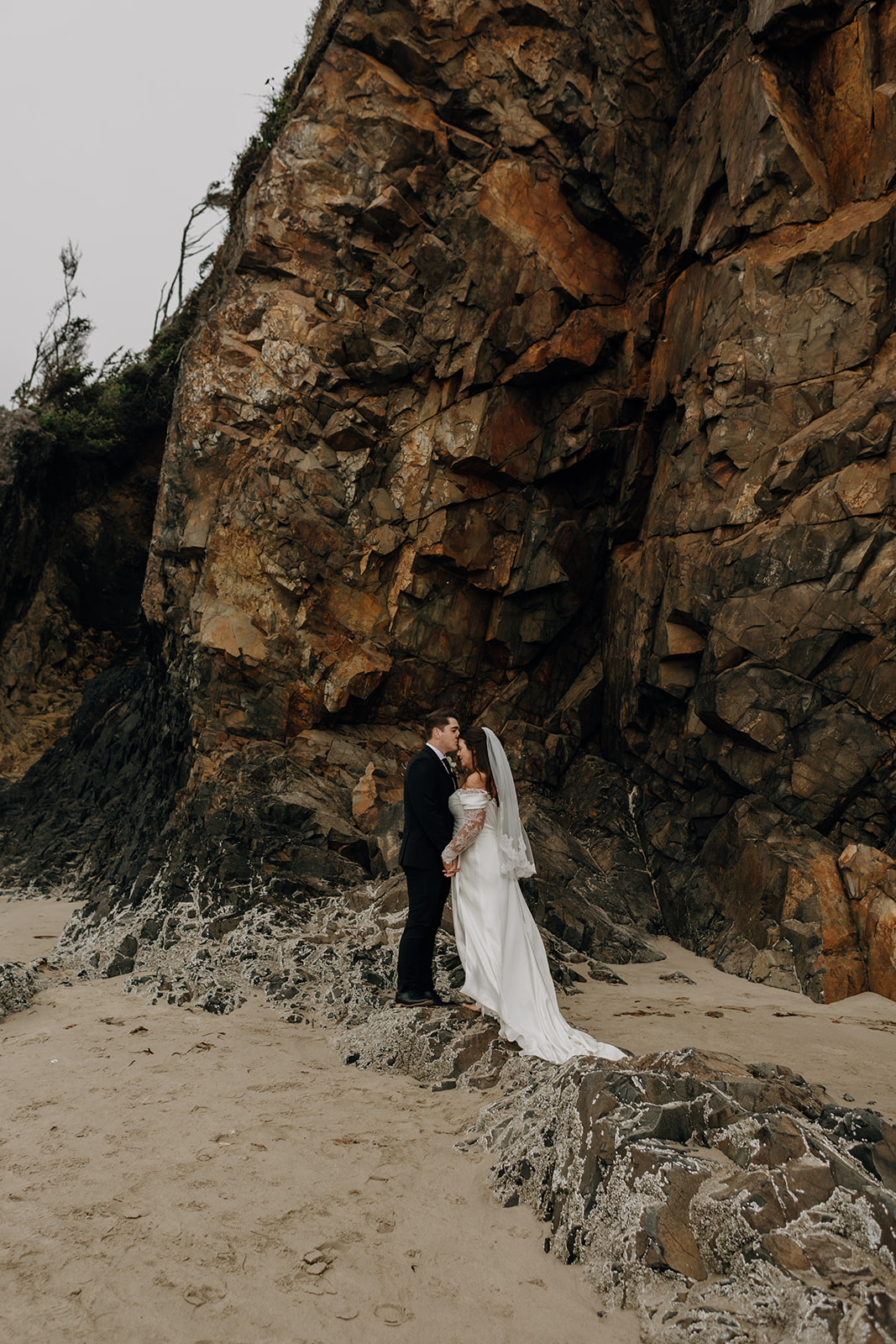 Bride and groom embrace on a sandy beach against a towering rocky cliff. The scene conveys romance and natural beauty under a cloudy sky.