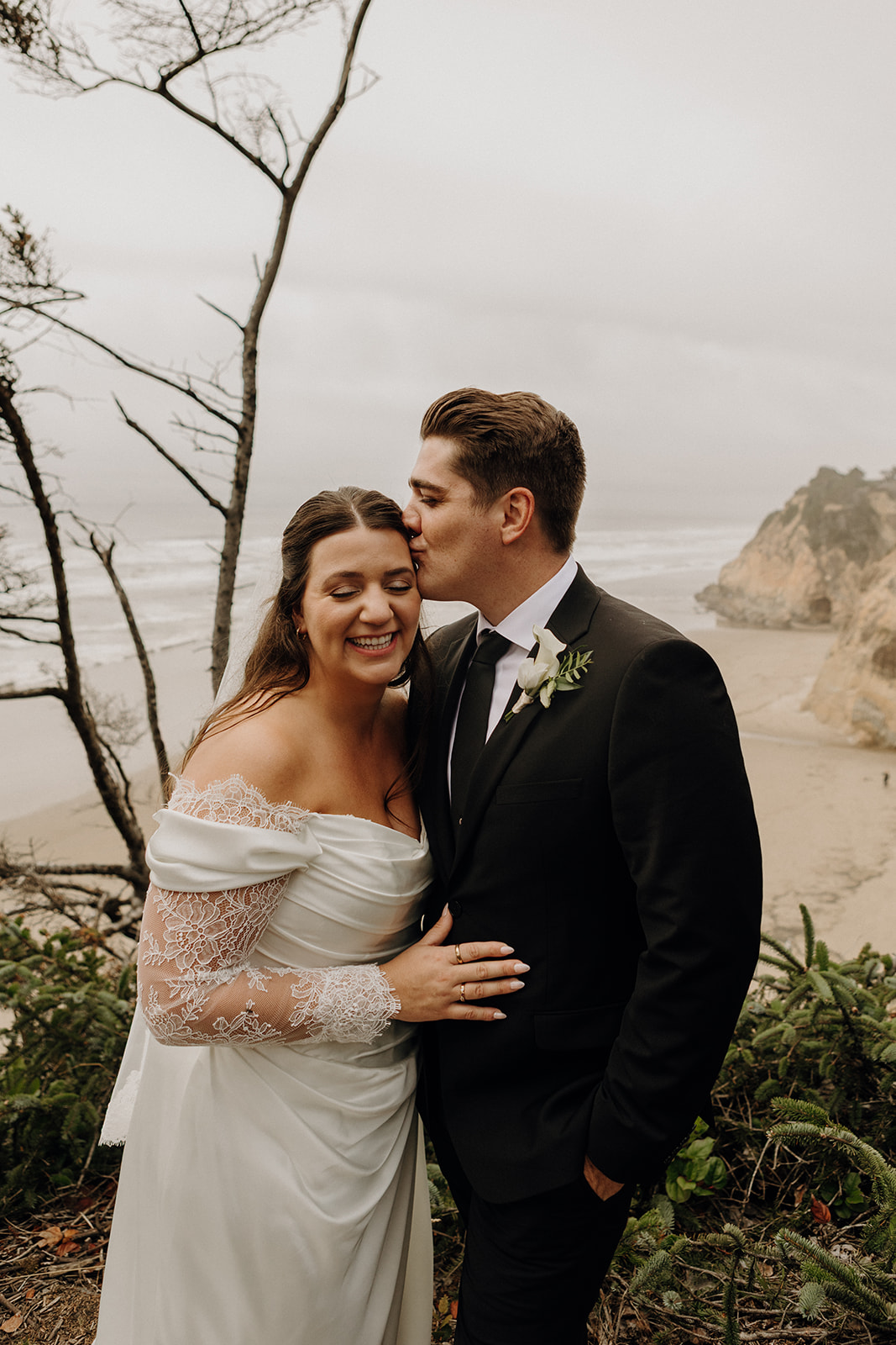 Bride and groom in wedding attire smiling joyfully by a beach. The groom kisses the bride's forehead, surrounded by trees under a cloudy sky.