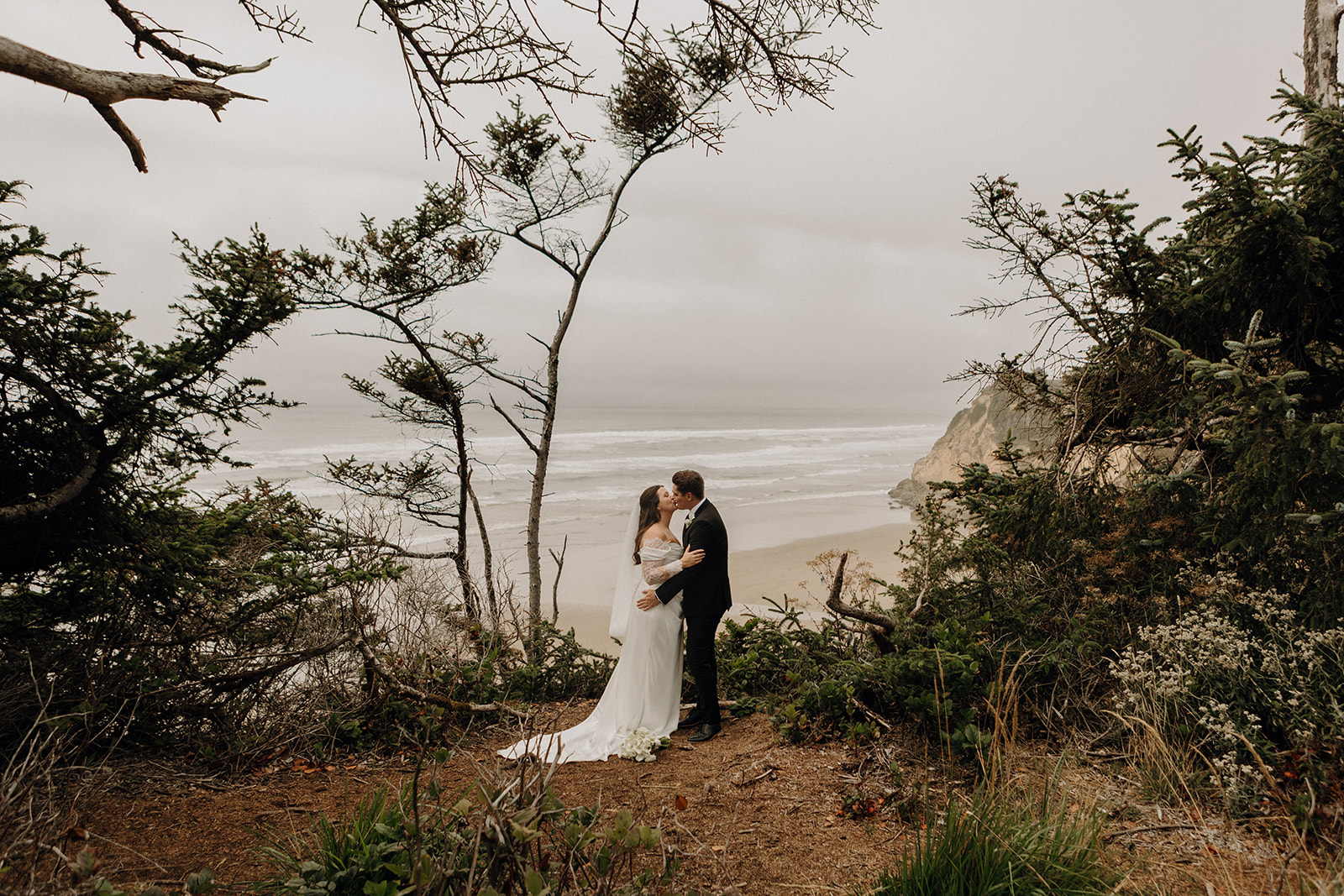 A couple in wedding attire embraces on a cliff overlooking a misty beach. Surrounded by lush greenery, the scene conveys romance and tranquility.