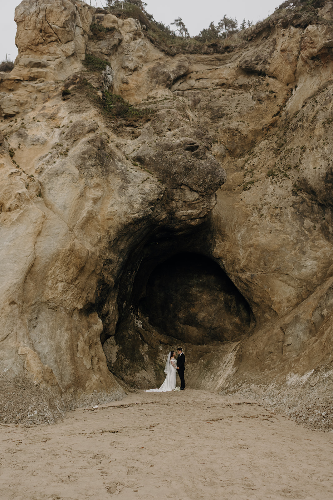 A couple in wedding attire stands at the entrance of a large, rugged cave on a sandy beach. The scene is dramatic and romantic.