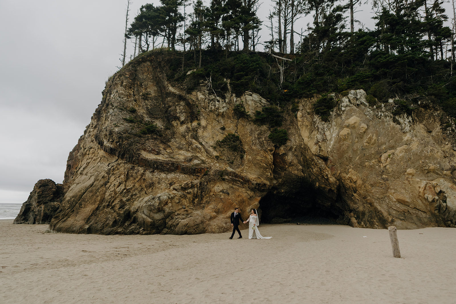 A couple walks hand in hand on a sandy beach with a dramatic, rocky cliff and dense greenery in the background. The scene is serene and overcast.