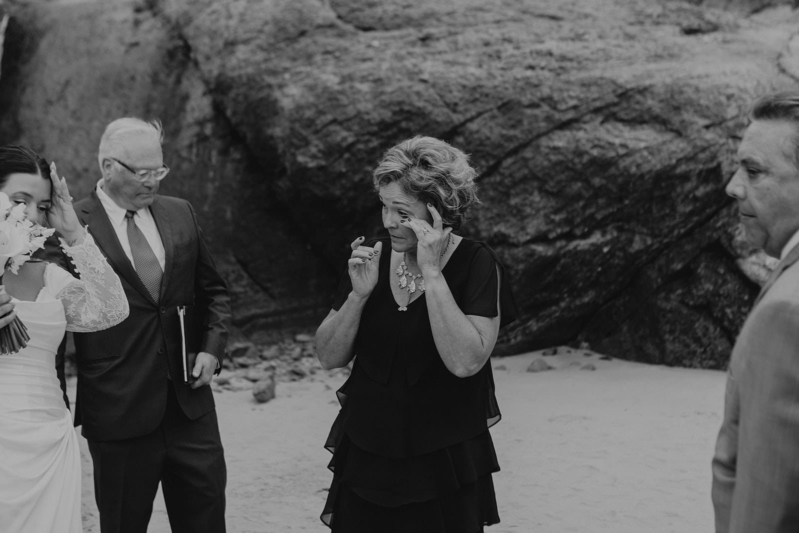 A black-and-white photo of a touching moment at a beach wedding. A woman in formal wear wipes away tears, while others look on near rocky cliffs.