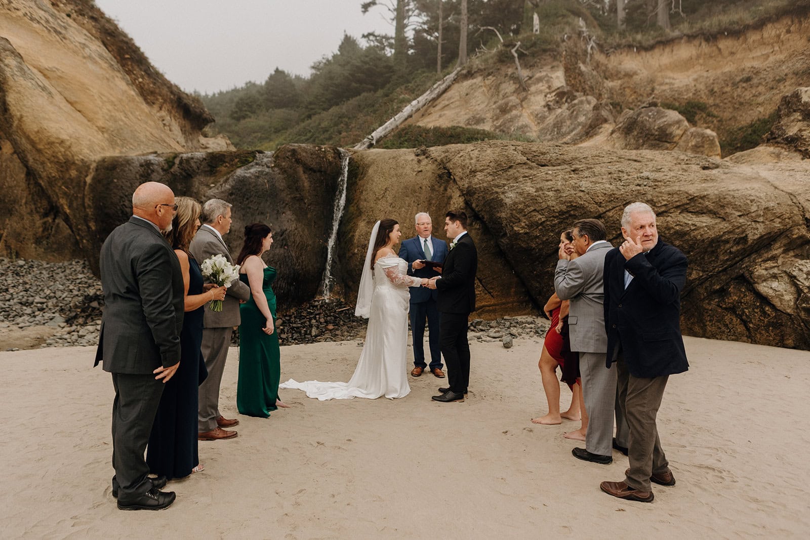 A beach wedding with a couple exchanging vows near a rocky cliff. A small group of guests in formal attire surrounds them, creating an intimate atmosphere.