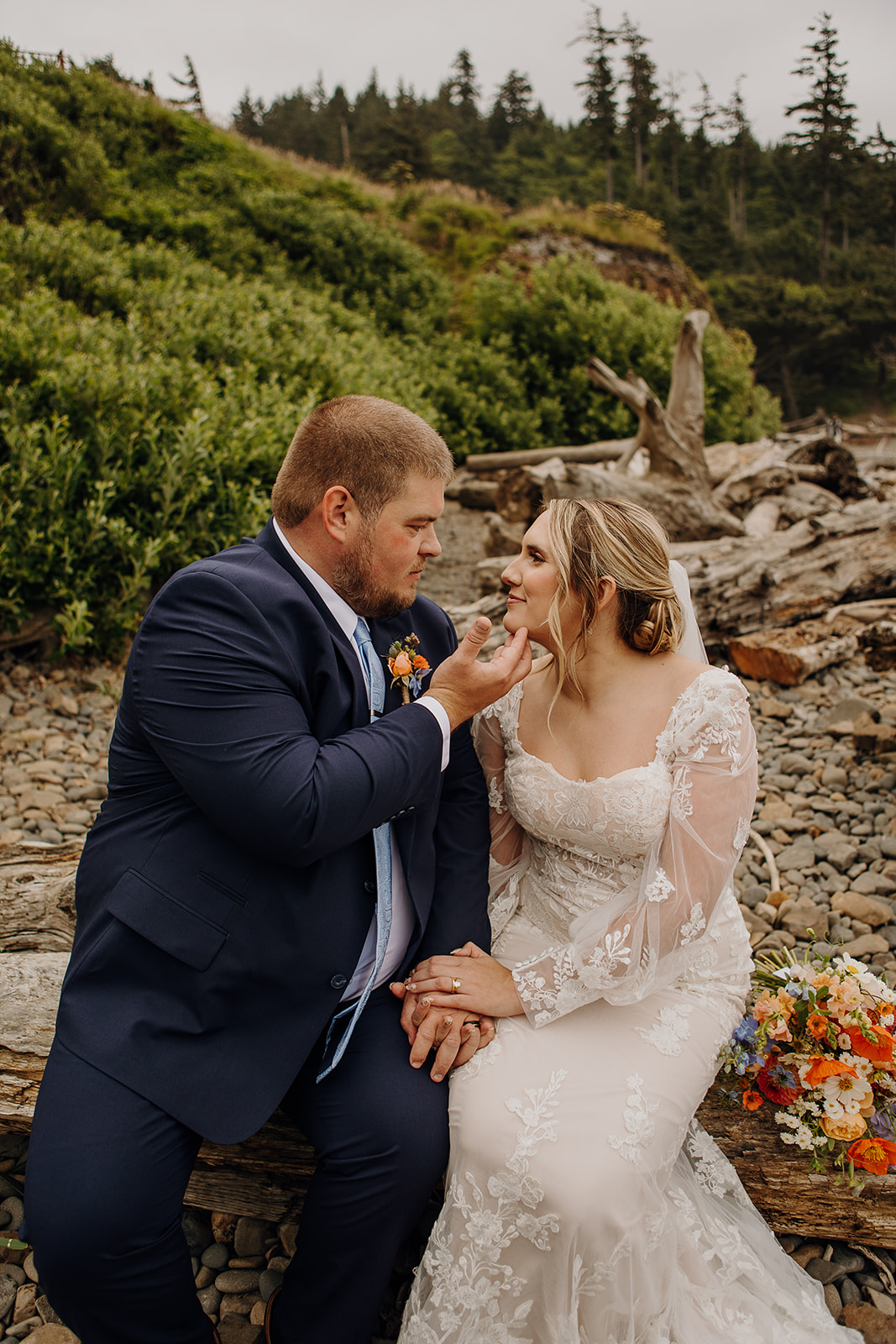 A couple sits on driftwood at a rocky beach. The groom in a navy suit gently touches the bride's chin. She wears a lace gown. A bridal bouquet and lush greenery are in the background.