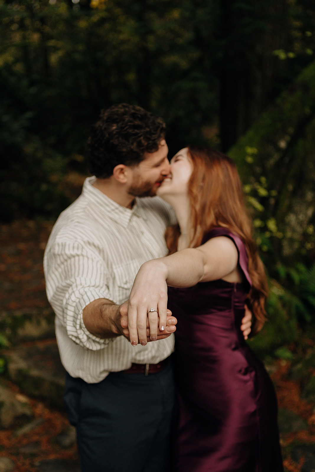 man and woman kiss in the background as they both hold out their held hands with the woman showing off her engagement ring in the foreground