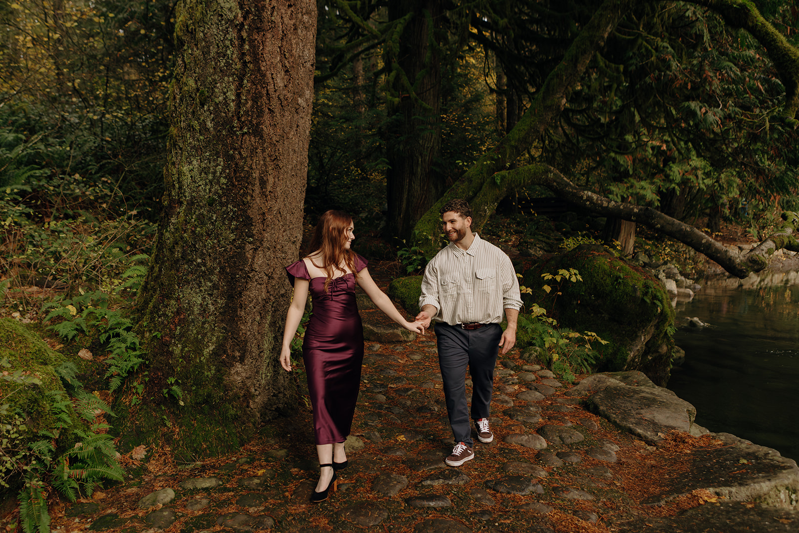 a young woman in a purple dress surrounded by lush green forest leads a young man by the hand both smiling