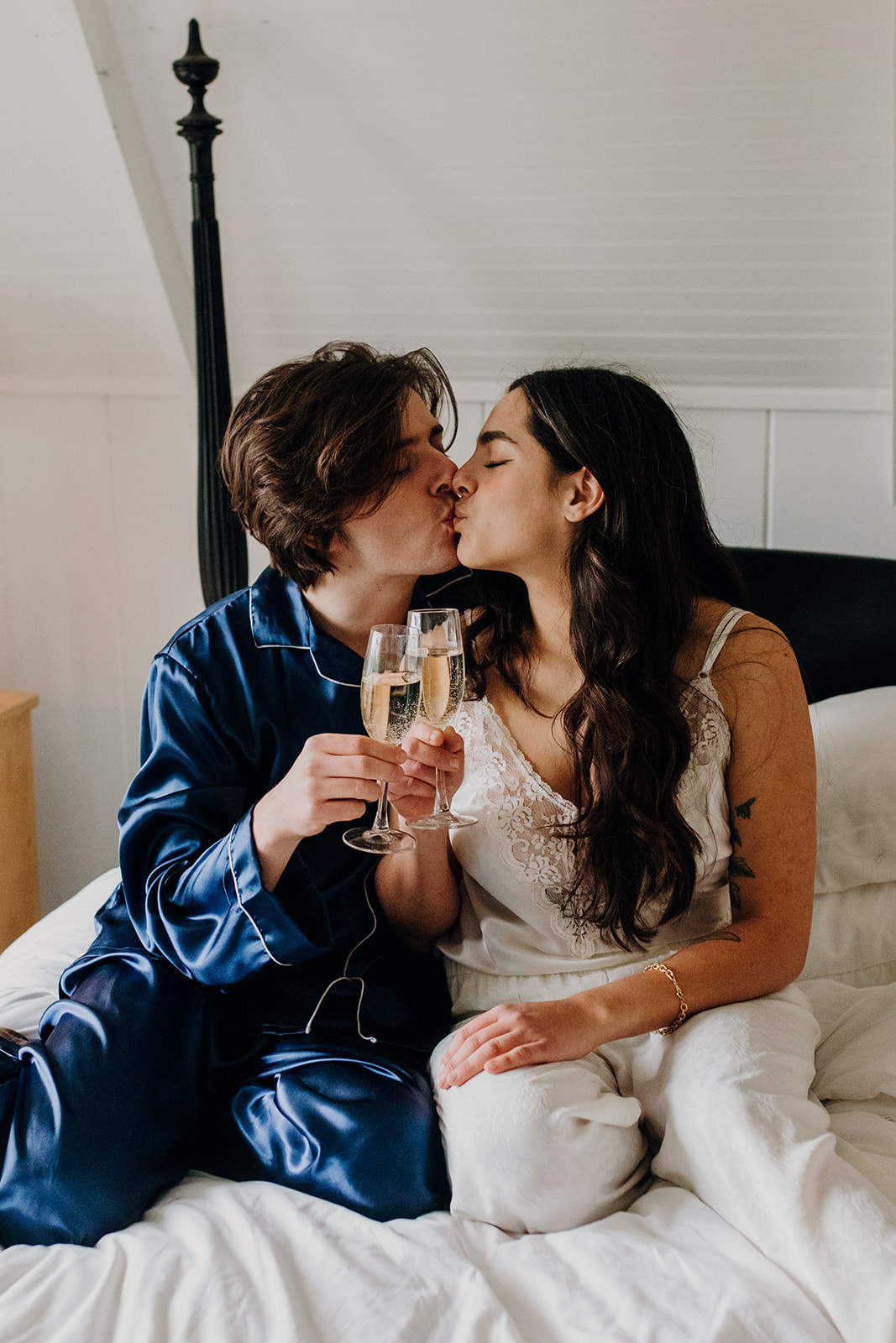 A couple in pajamas kisses on a bed, holding champagne glasses. The atmosphere is intimate and celebratory, set in a cozy, softly lit room.