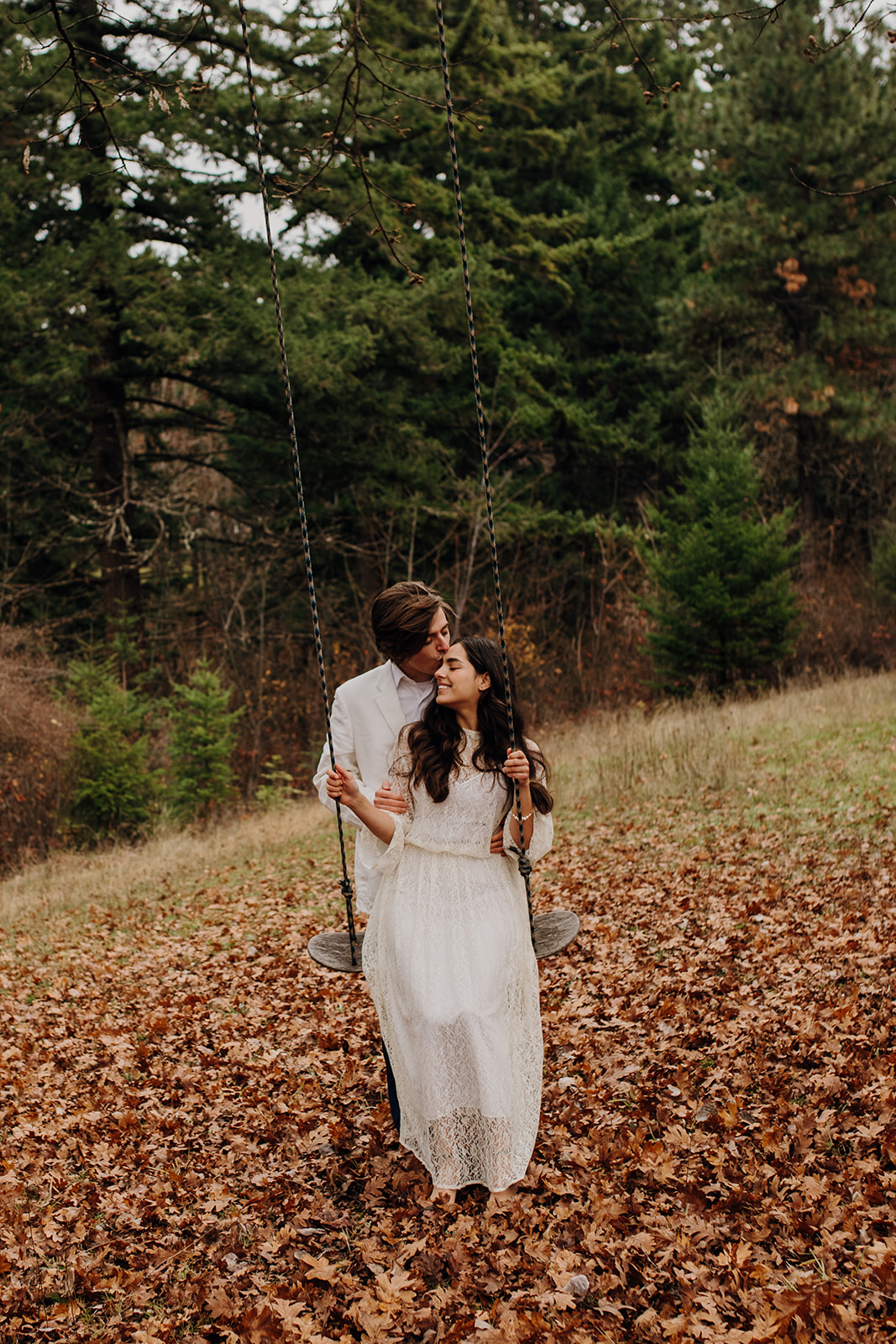 A couple in elegant attire shares a tender moment on a swing amid a forest backdrop. The ground is covered in autumn leaves, exuding a romantic, serene vibe.