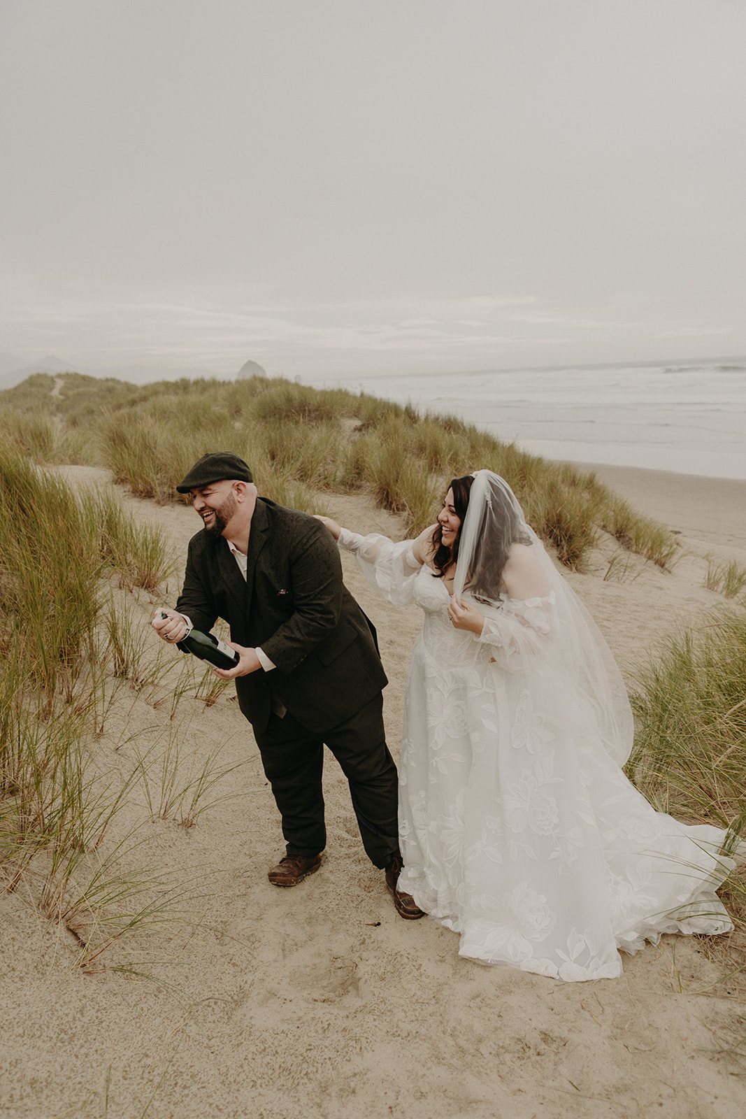 bride and groom laugh together on a beach as they uncork a champagne bottle