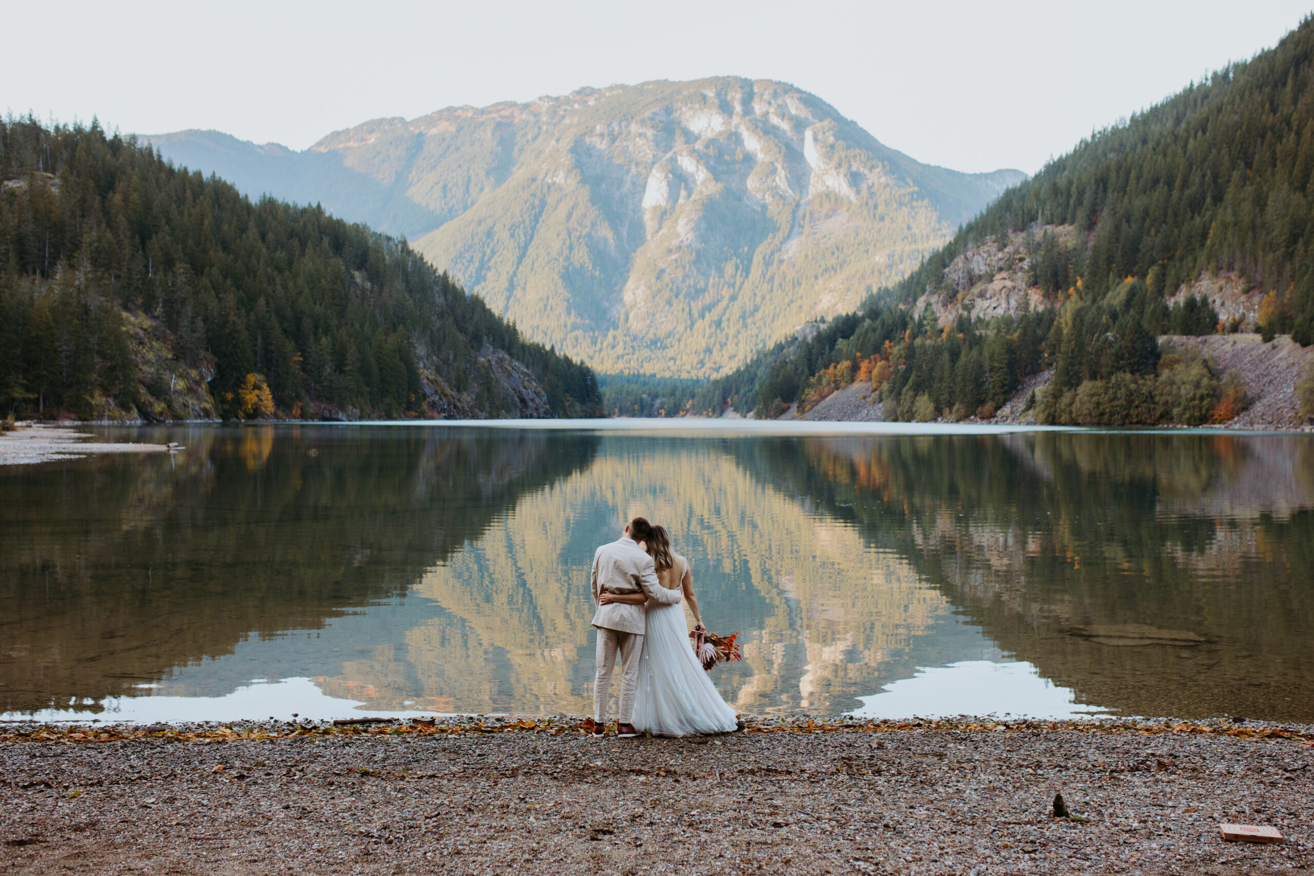man and woman stand on the shores of Diablo Lake in wedding attire looking towards the mountain reflected in the lake's water with their backs to the camera