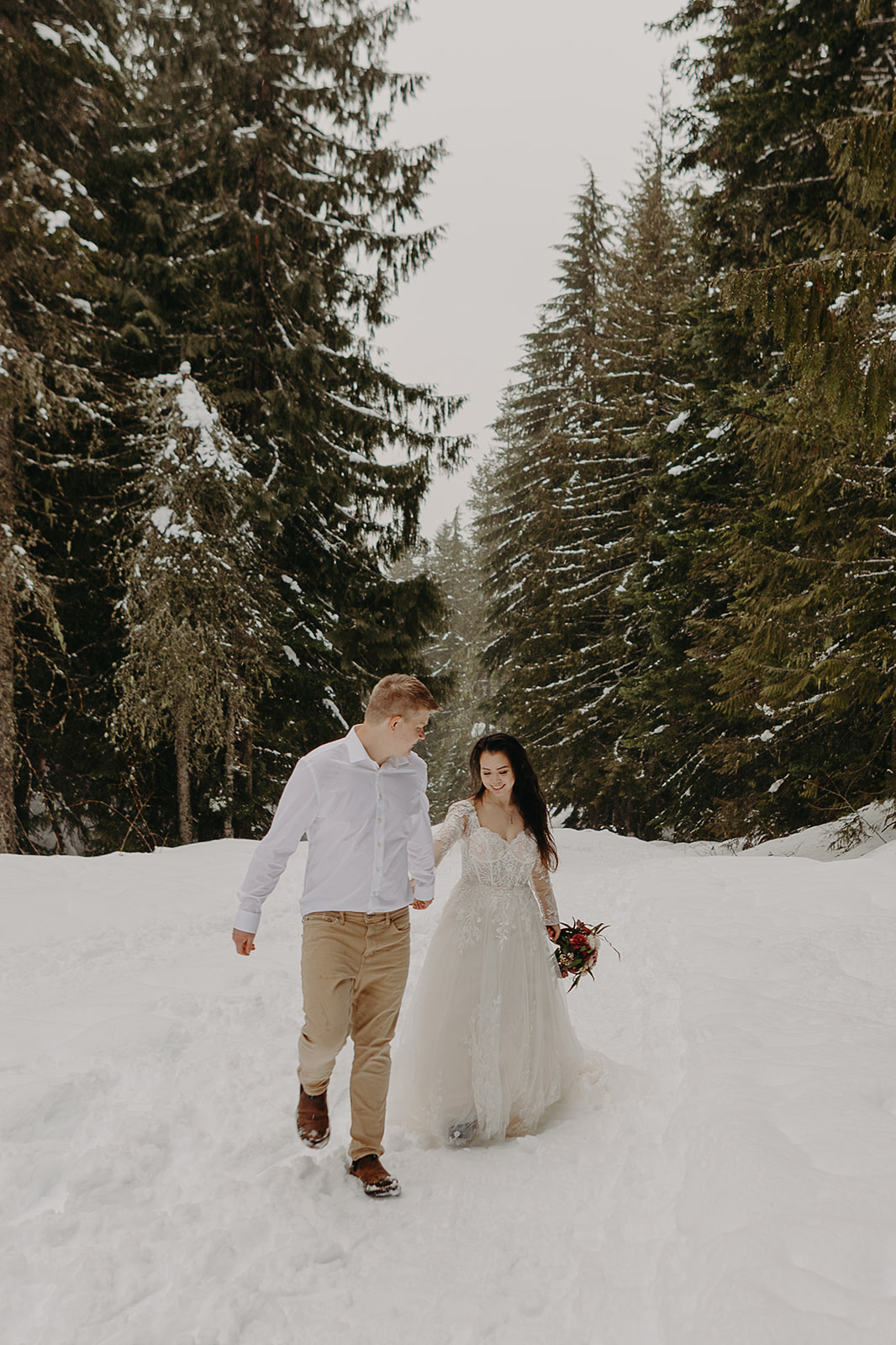 man and woman in wedding dress hold hands and walk through snow in an evergreen forest