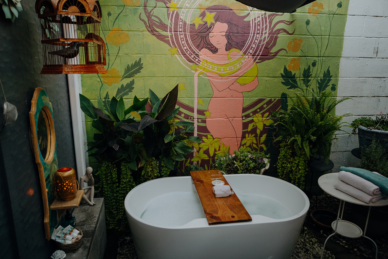 A serene outdoor bathroom features a filled white tub with a wooden tray, surrounded by lush plants. A colorful mural of a woman adorns the wall, conveying tranquility.