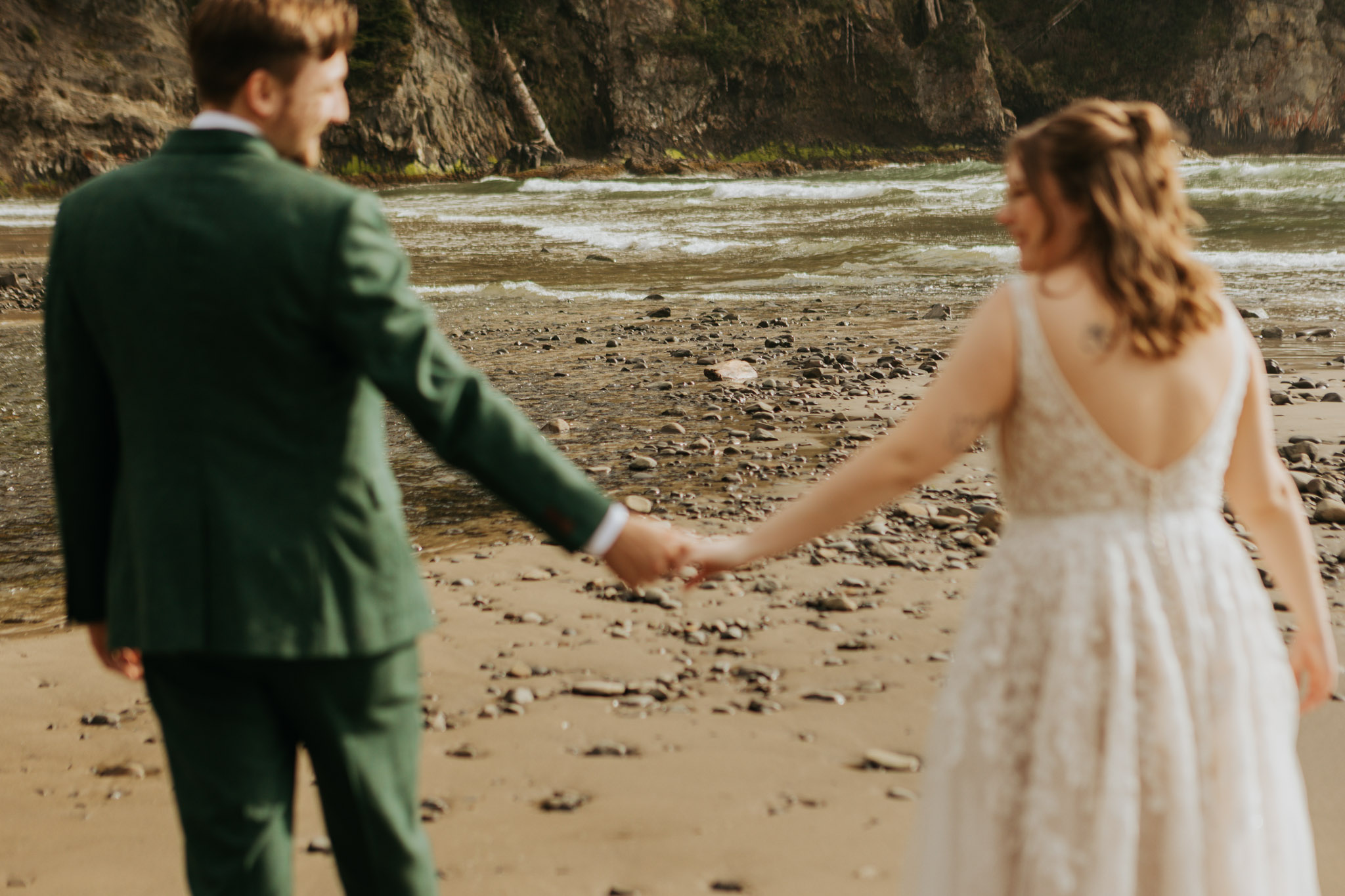 A couple walks hand in hand on a rocky beach. The man wears a green suit, and the woman is in a white dress. The scene conveys romance and tranquility.