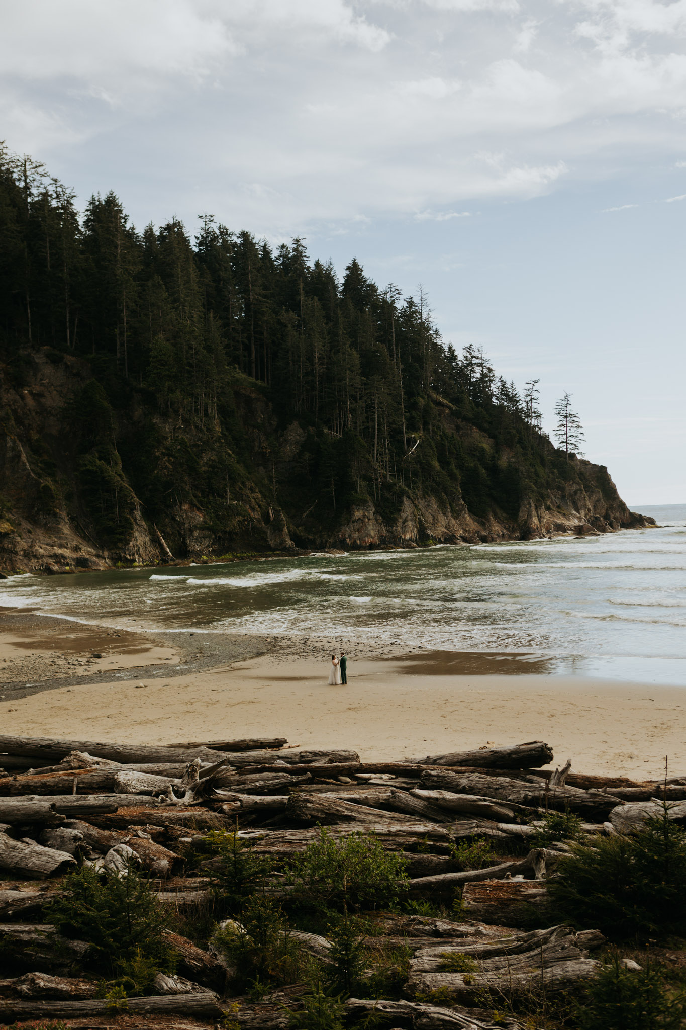 A serene coastal scene with a couple standing on a sandy beach, surrounded by driftwood and lush fir trees on a rocky cliffside. A peaceful, natural atmosphere.
