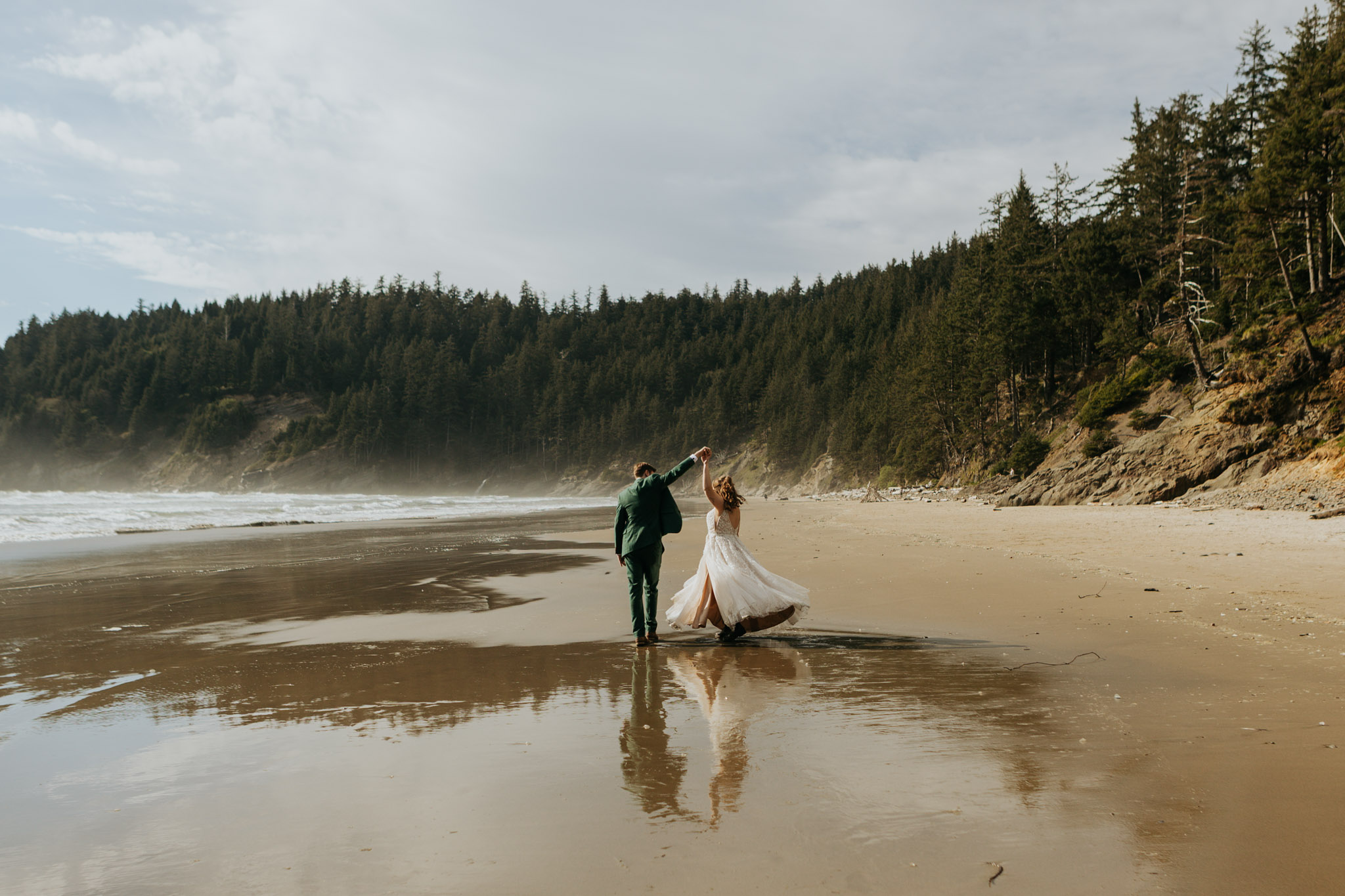 A couple dances joyfully on a serene beach, surrounded by forested cliffs. The bride's dress flows elegantly, reflecting in the wet sand.