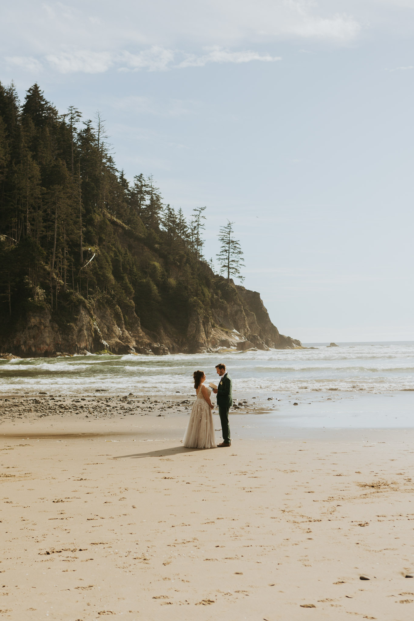 A couple stands on a sandy beach, holding hands under a clear blue sky. A rocky, forested cliff rises to the left, with gentle ocean waves in the background. Peaceful and romantic.