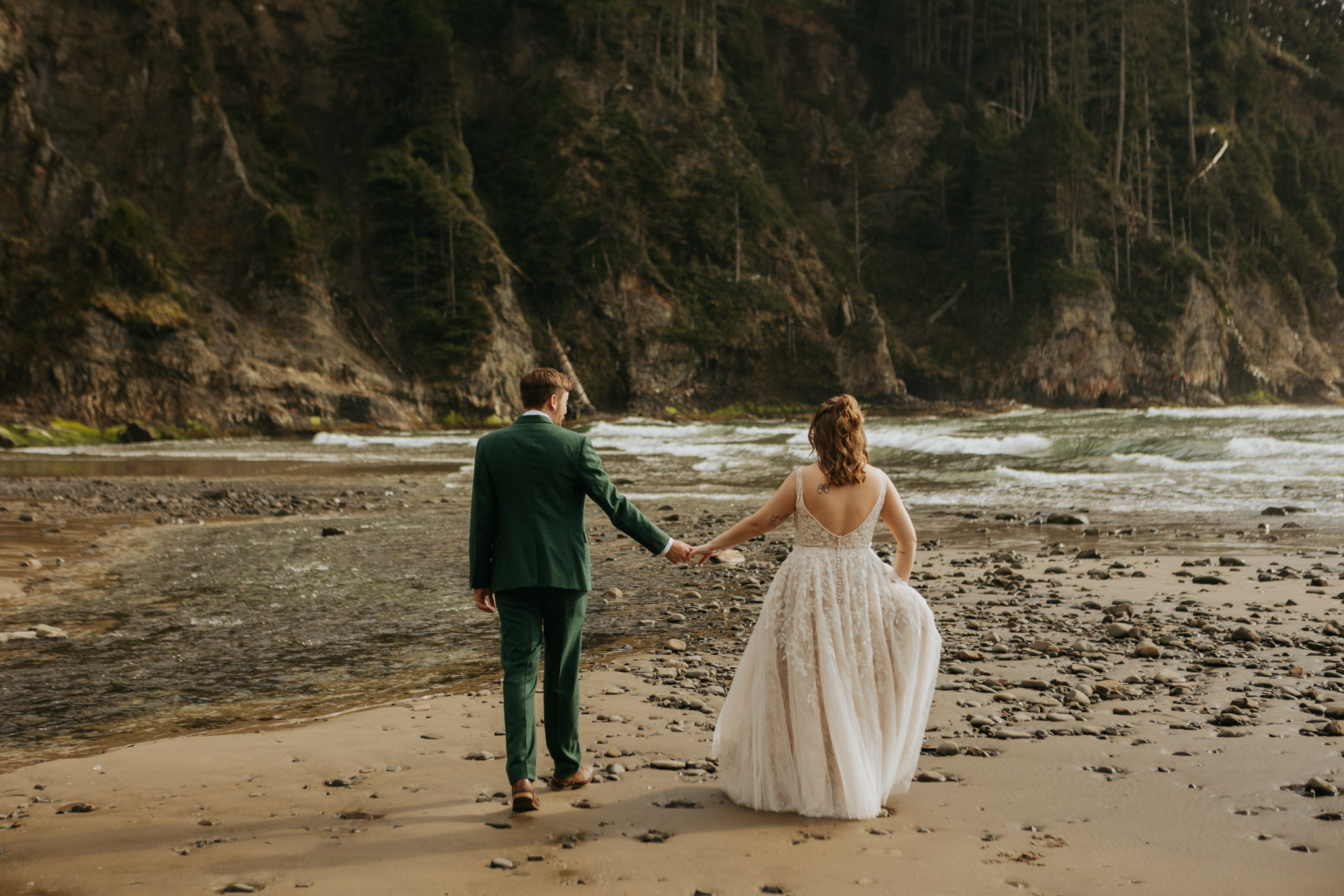 A couple in formal attire walks hand in hand on a rocky beach. The woman wears a flowing white dress, and the man dons a green suit. A tranquil, scenic coastline lies ahead.