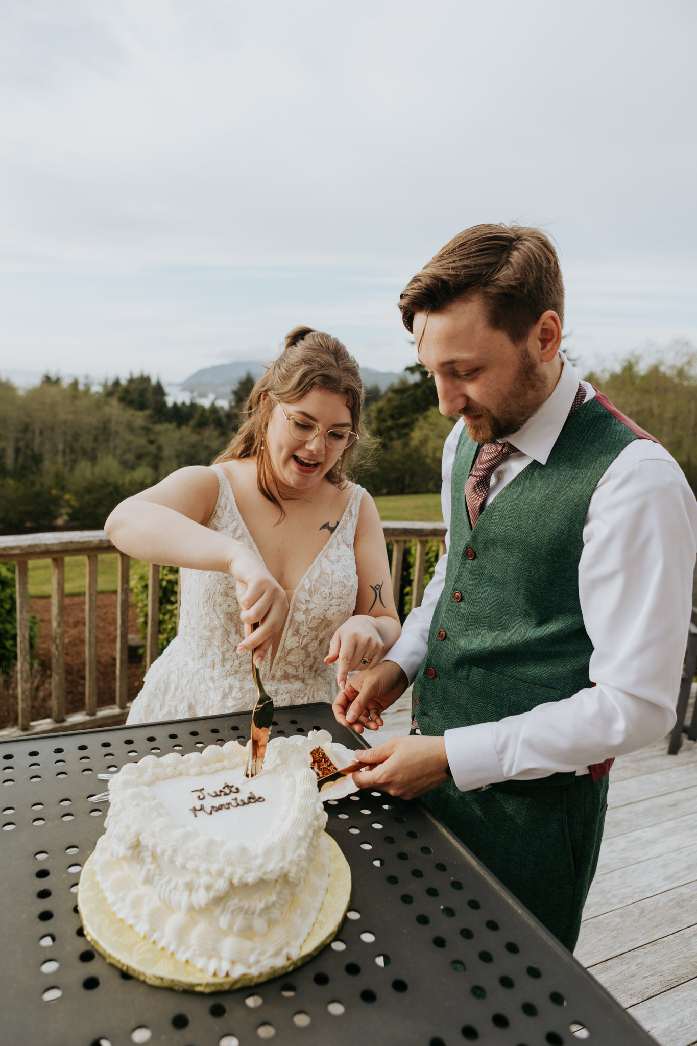 A couple in wedding attire joyfully cuts a white cake on a patio. The scene is lighthearted with a scenic outdoor backdrop of trees and sky.