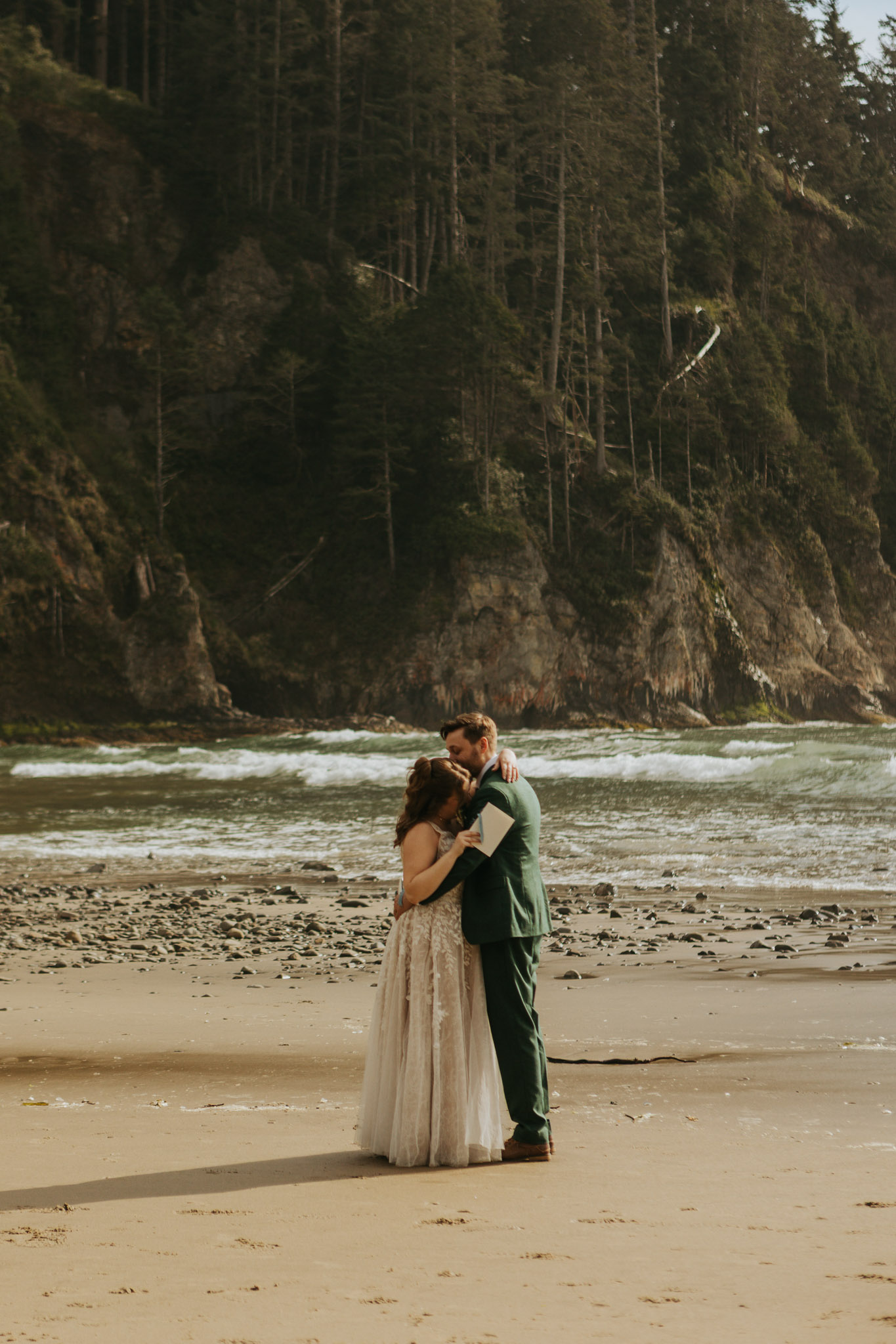 A couple embraces on a rocky beach, the woman in a white dress and the man in a green suit. They stand before a backdrop of cliffs and ocean waves.