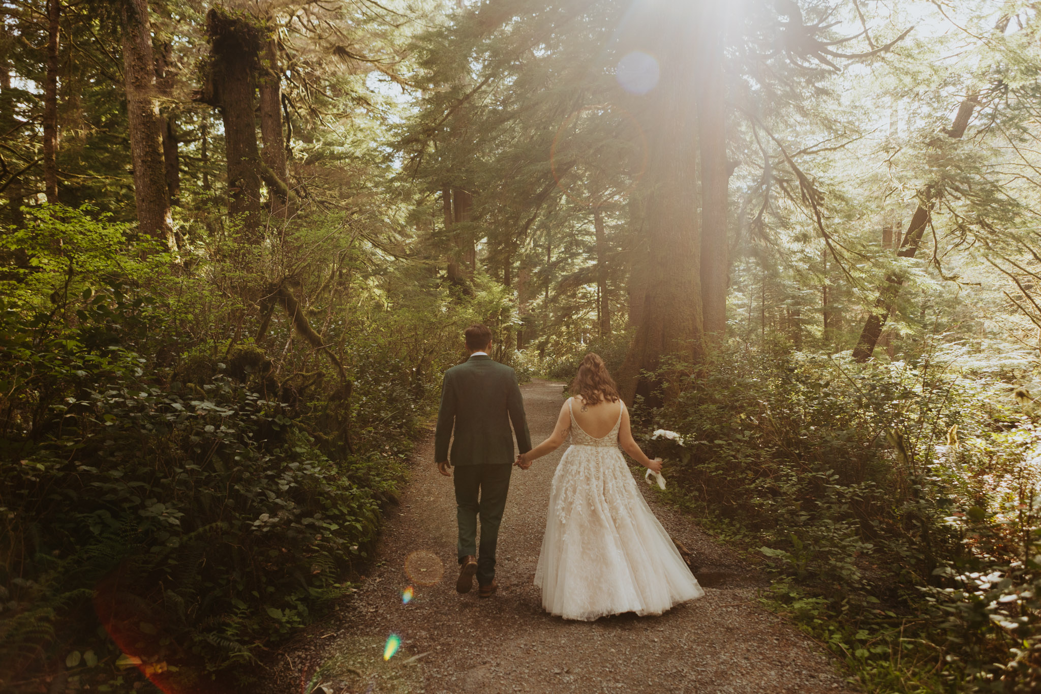 A couple walks hand in hand down a forest path, surrounded by lush green trees. Sunlight streams through the branches, creating a peaceful, romantic atmosphere.