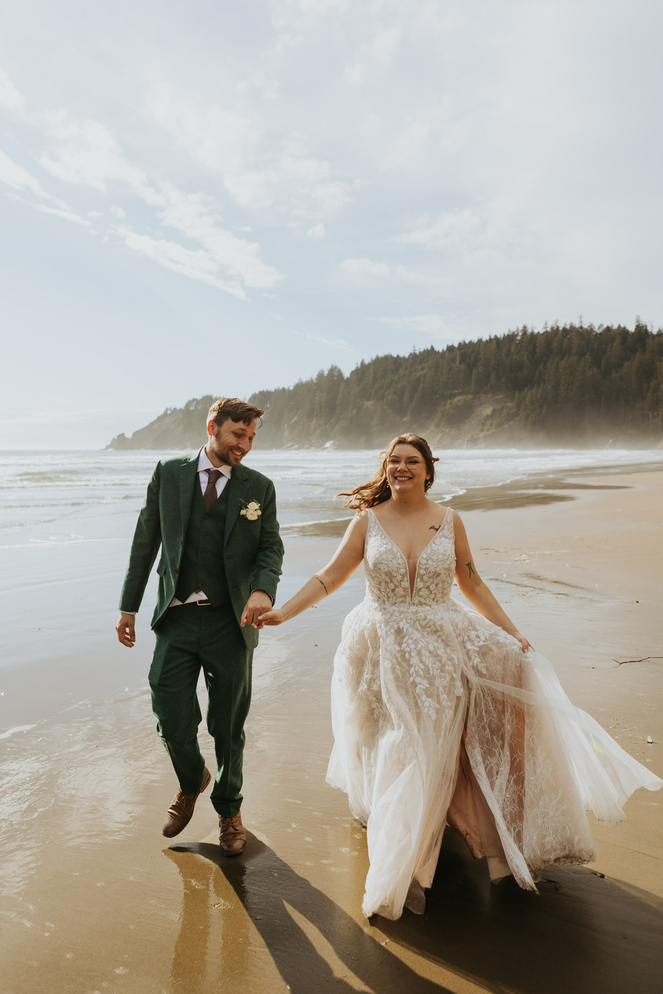 A joyful couple in wedding attire walks hand-in-hand along a sandy beach, with gentle waves and a forested cliff in the background under a bright sky.