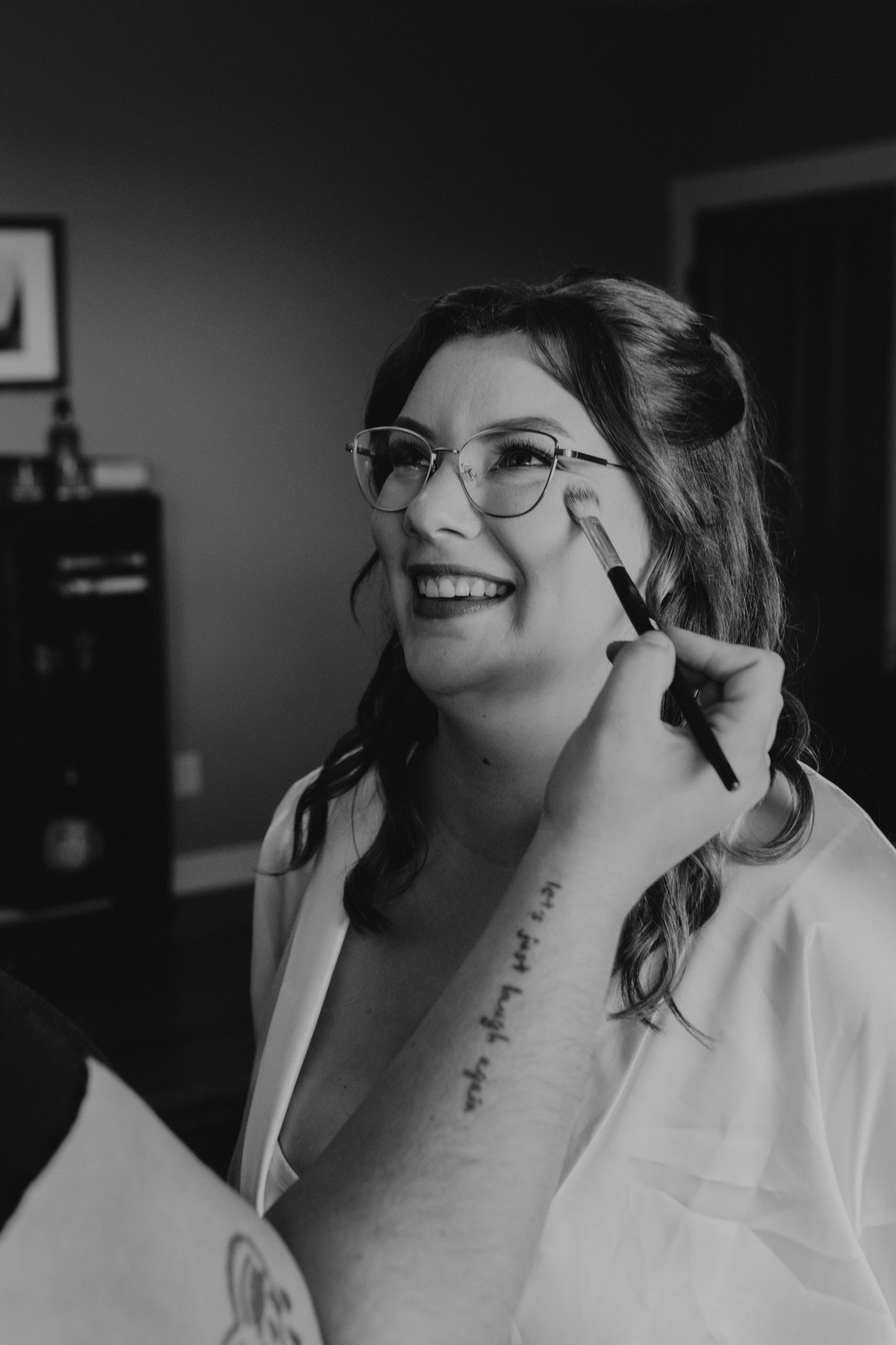A woman with glasses in a wedding robe smiles as someone applies makeup with a brush on her cheek. The black-and-white image exudes joy and anticipation in a calm setting.