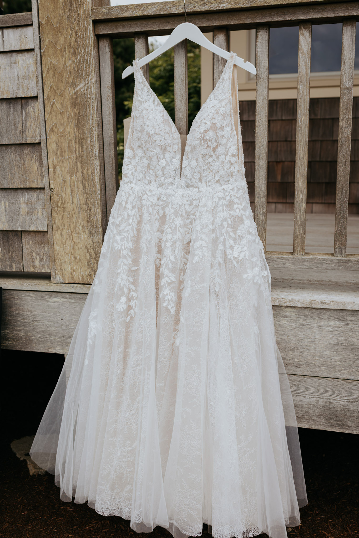 Elegant white lace wedding dress on a hanger, displayed against rustic wooden siding. The dress has a deep V-neck and intricate embroidery.