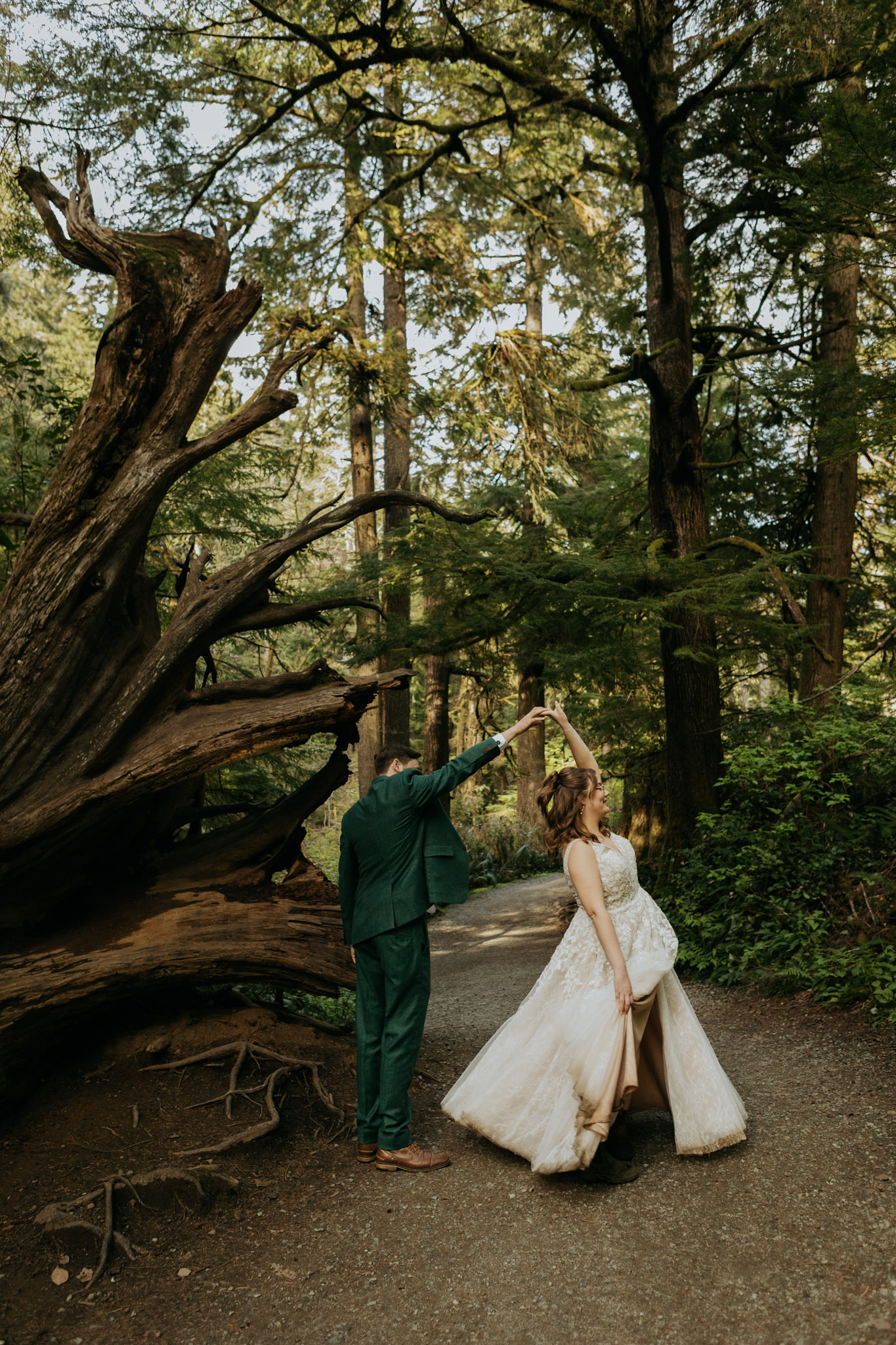 A couple dances joyfully in a forest. The groom in a green suit twirls the bride in a white dress. A fallen tree adds rustic charm to the lush, green setting.