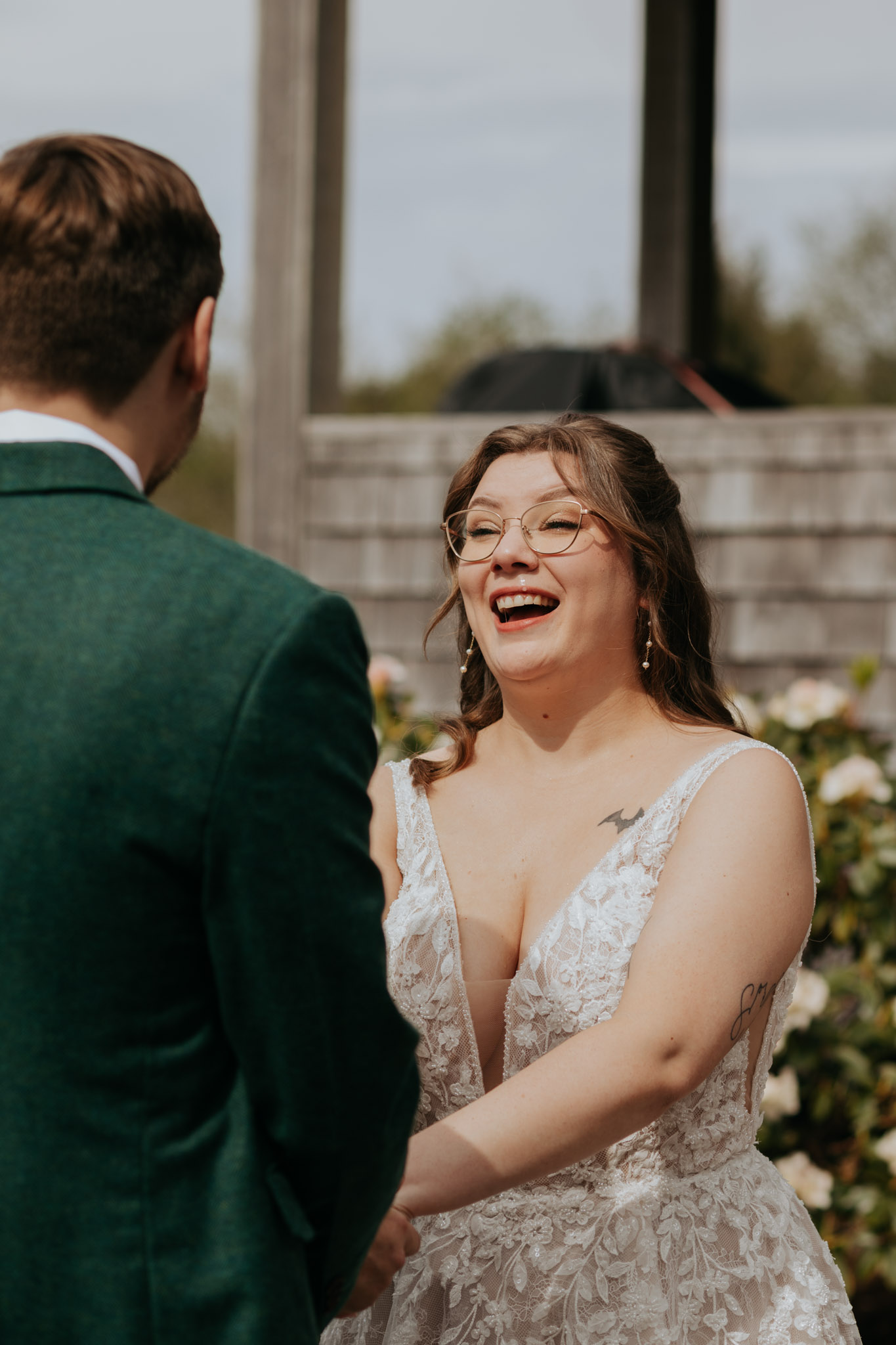A bride in a lace wedding dress and glasses laughs joyfully while holding hands with a man in a green suit, outdoors with a soft-focus floral background.