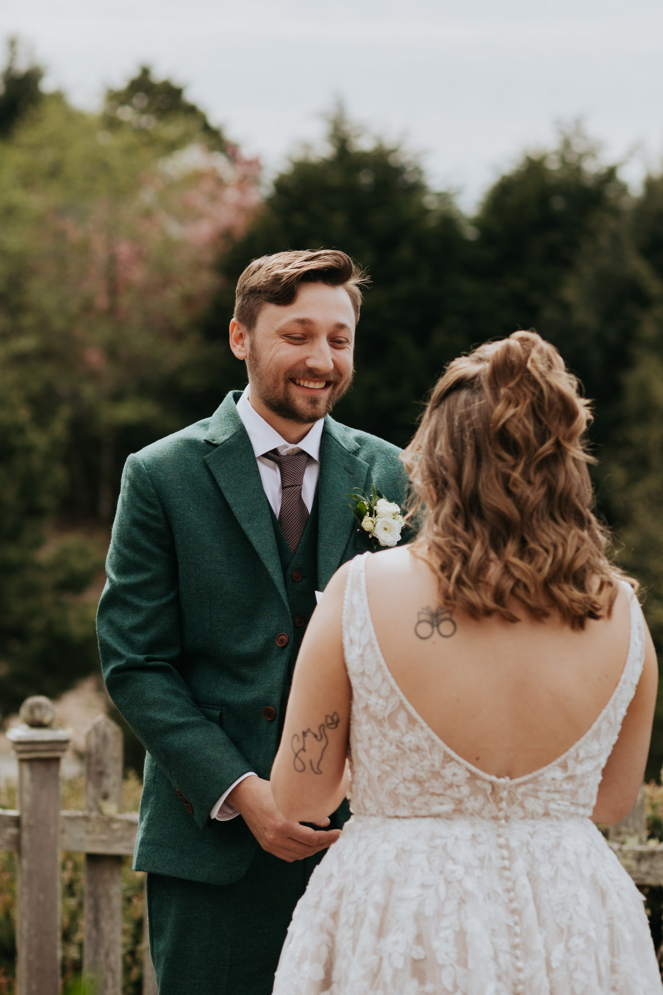 A groom in a green suit and smiling bride in a lace gown hold hands outdoors, surrounded by green trees, creating a joyful, intimate wedding moment.