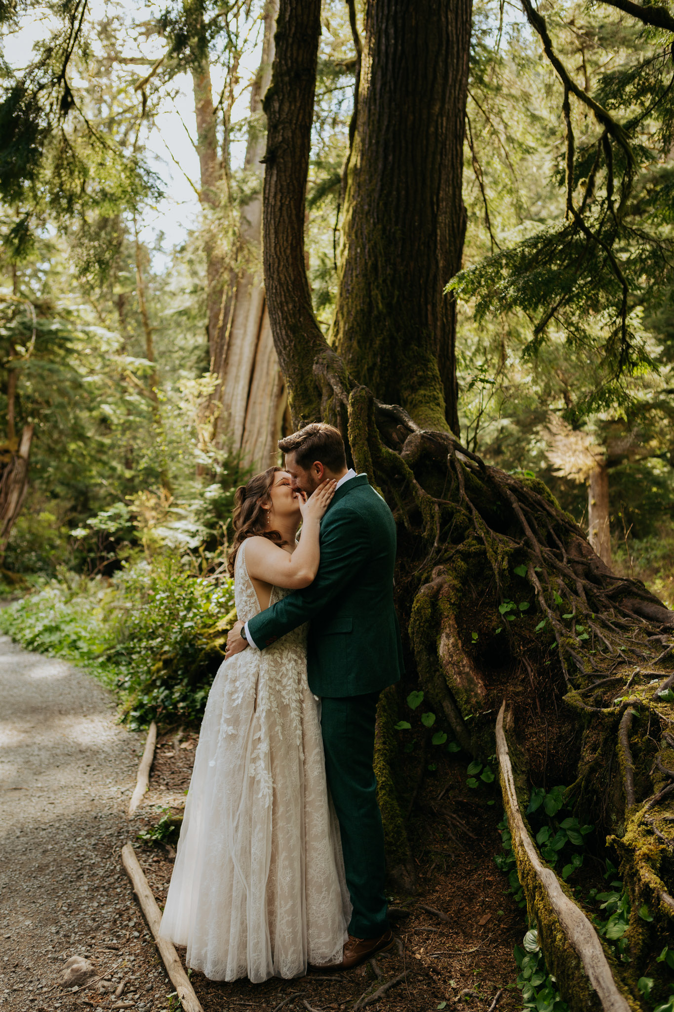 Bride and groom embrace lovingly under a towering tree in a lush forest, conveying romance and tranquility on a sunlit trail.