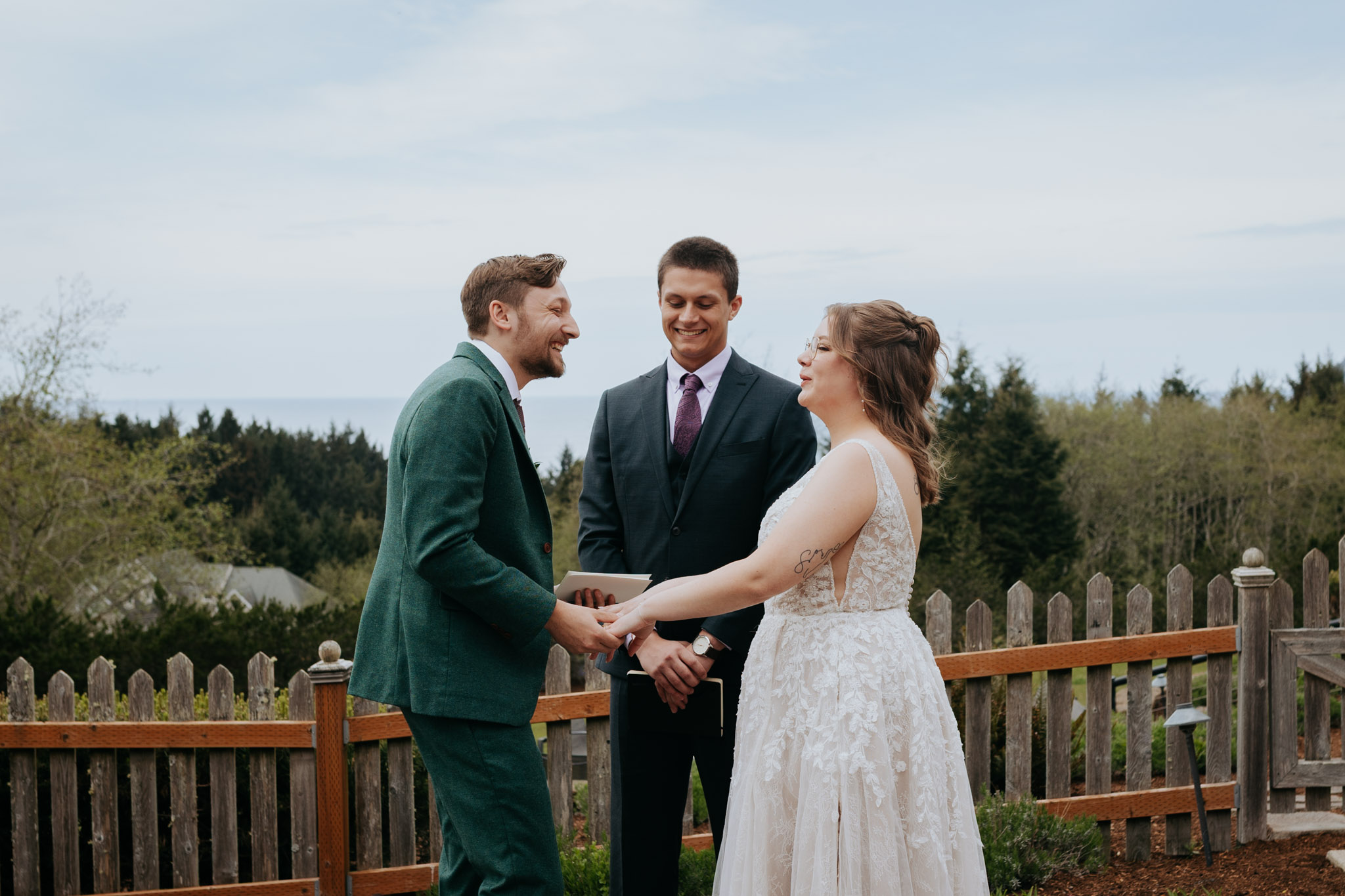 Bride and groom hold hands, smiling at each other during an outdoor wedding ceremony. An officiant stands behind them. Trees and a fence are in the background.