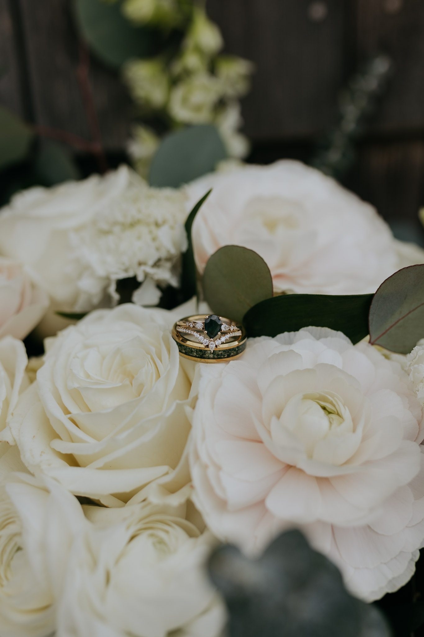 Elegant green wedding rings rest on a bouquet of white roses and peonies, conveying a romantic and serene mood against a blurred natural background.